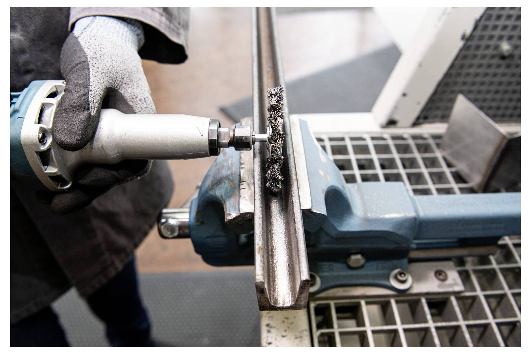 „Eine Person benutzt ein Elektrowerkzeug, um eine Metallschiene zu reinigen, die in einem blauen Schraubstock eingespannt ist und dabei Wartungs- oder Bauarbeiten demonstriert.