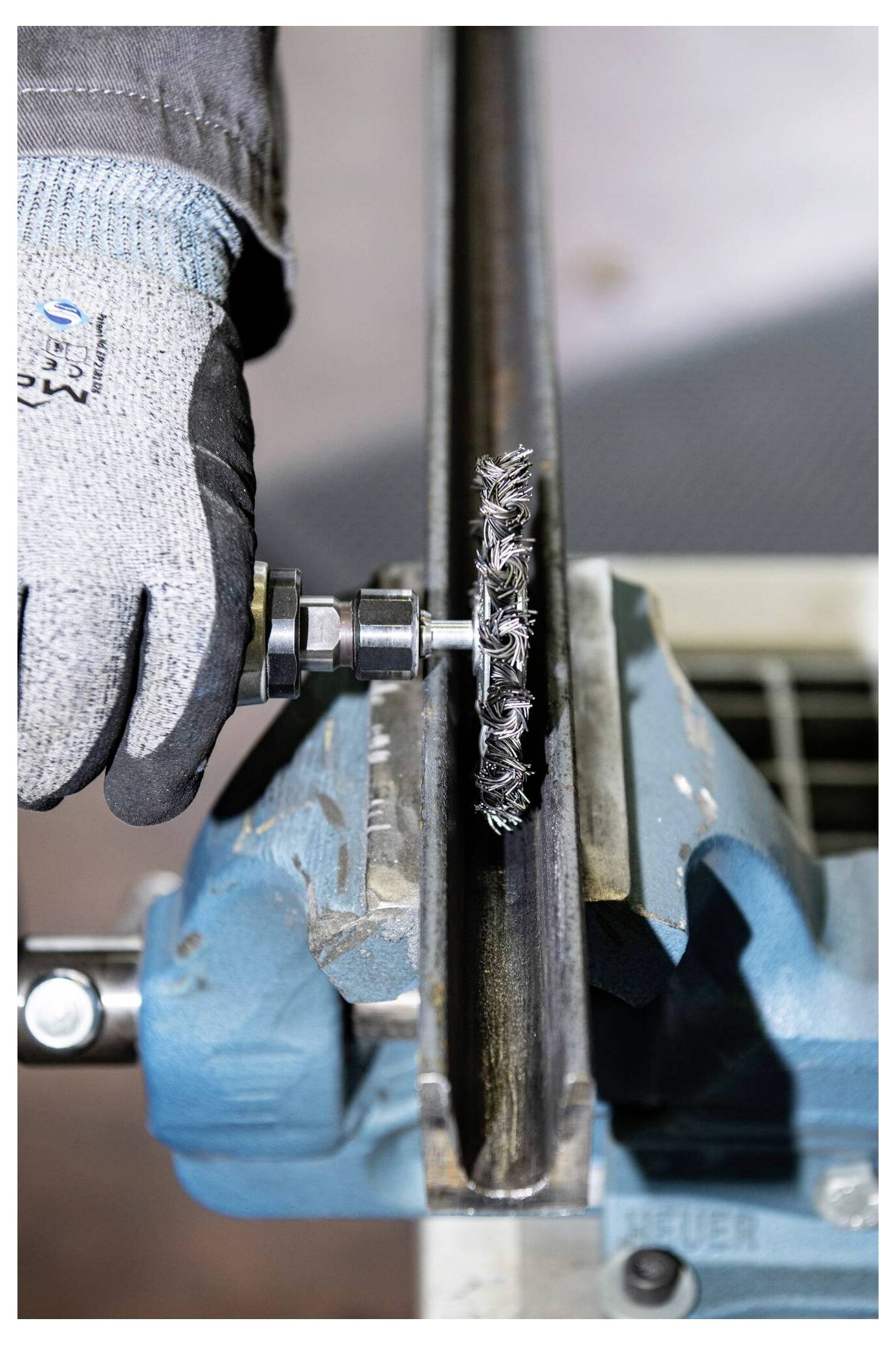 Eine Person mit einem Schutzhandschuh bedient ein Elektrowerkzeug mit einer Drahtbürsten-Aufsatz, um eine in einer blauen Schraubzwinge gehaltene Metalloberfläche zu reinigen oder zu schleifen.