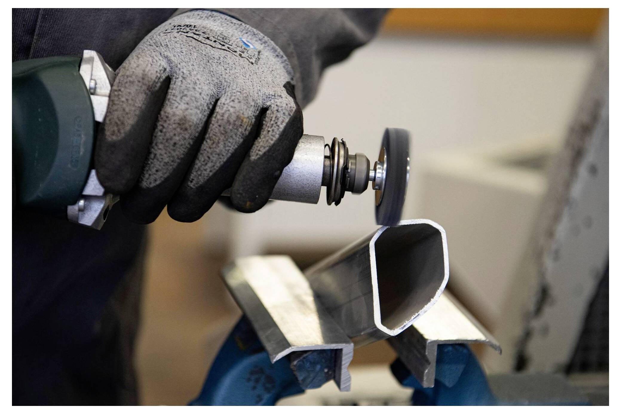 Eine Person mit Handschuhen verwendet ein Elektrowerkzeug, um ein Rohr aus Metall zu schneiden, das von einem Schraubstock fixiert wird, was auf industrielle oder Bauarbeiten hindeutet.