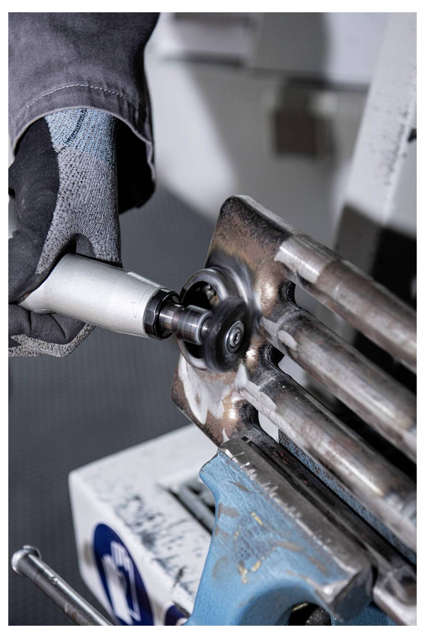 Eine Person mit Handschuhen verwendet ein Schleifwerkzeug an Metallstangen und glättet deren Oberfläche. Die Szene deutet auf eine industrielle oder metallverarbeitende Tätigkeit hin.