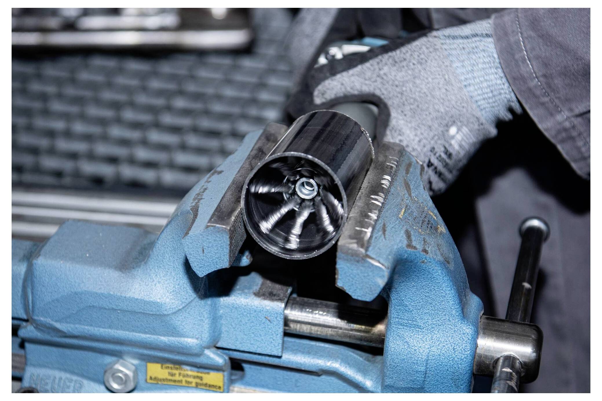 Eine Handschuh tragende Hand hält ein zylindrisches Metallobjekt mit einem komplexen Innendesign, das in einem blauen Schraubstock gesichert ist und präzise Arbeit in einer Werkstattumgebung andeutet.