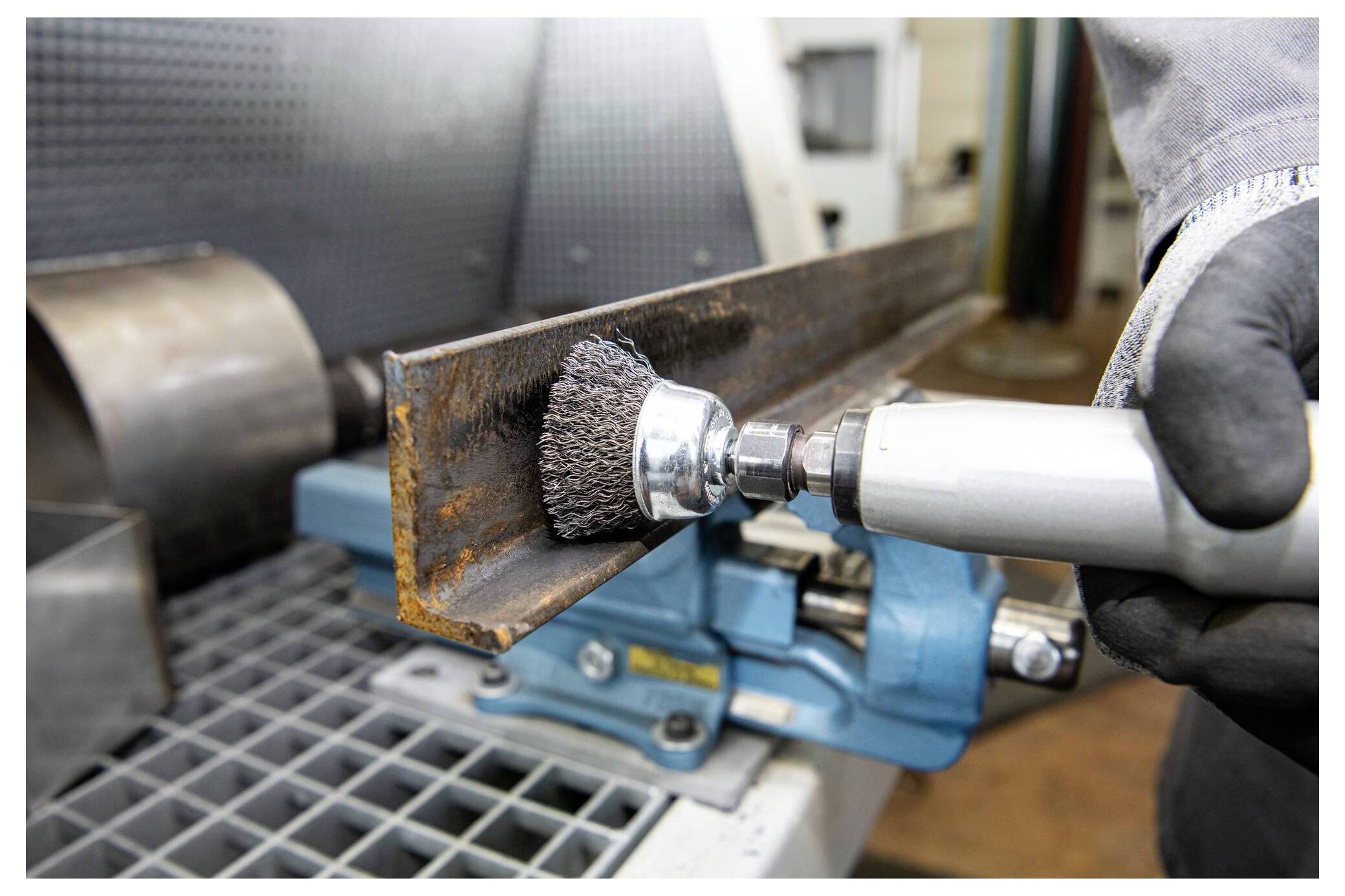 Ein Arbeiter verwendet einen Drahtbürstenaufsatz an einem Elektrowerkzeug, um Rost von einem Metallträger zu entfernen, der in einem blauen Schraubstock auf einer Werkbank befestigt ist.