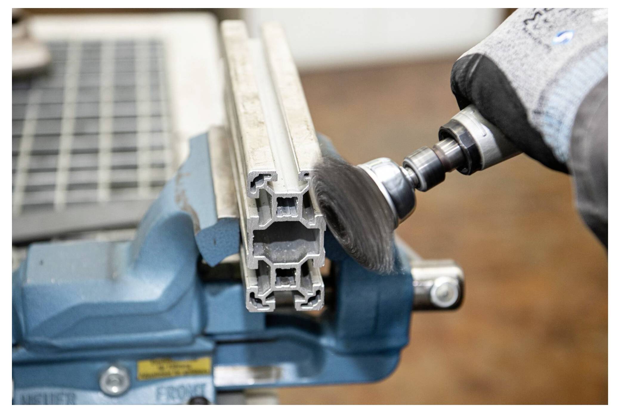 Ein Arbeiter poliert ein Metallstück, das in einem Schraubstock eingespannt ist, und verwendet dabei ein Rotationswerkzeug mit einer Drahtbürsten-Aufsatz.