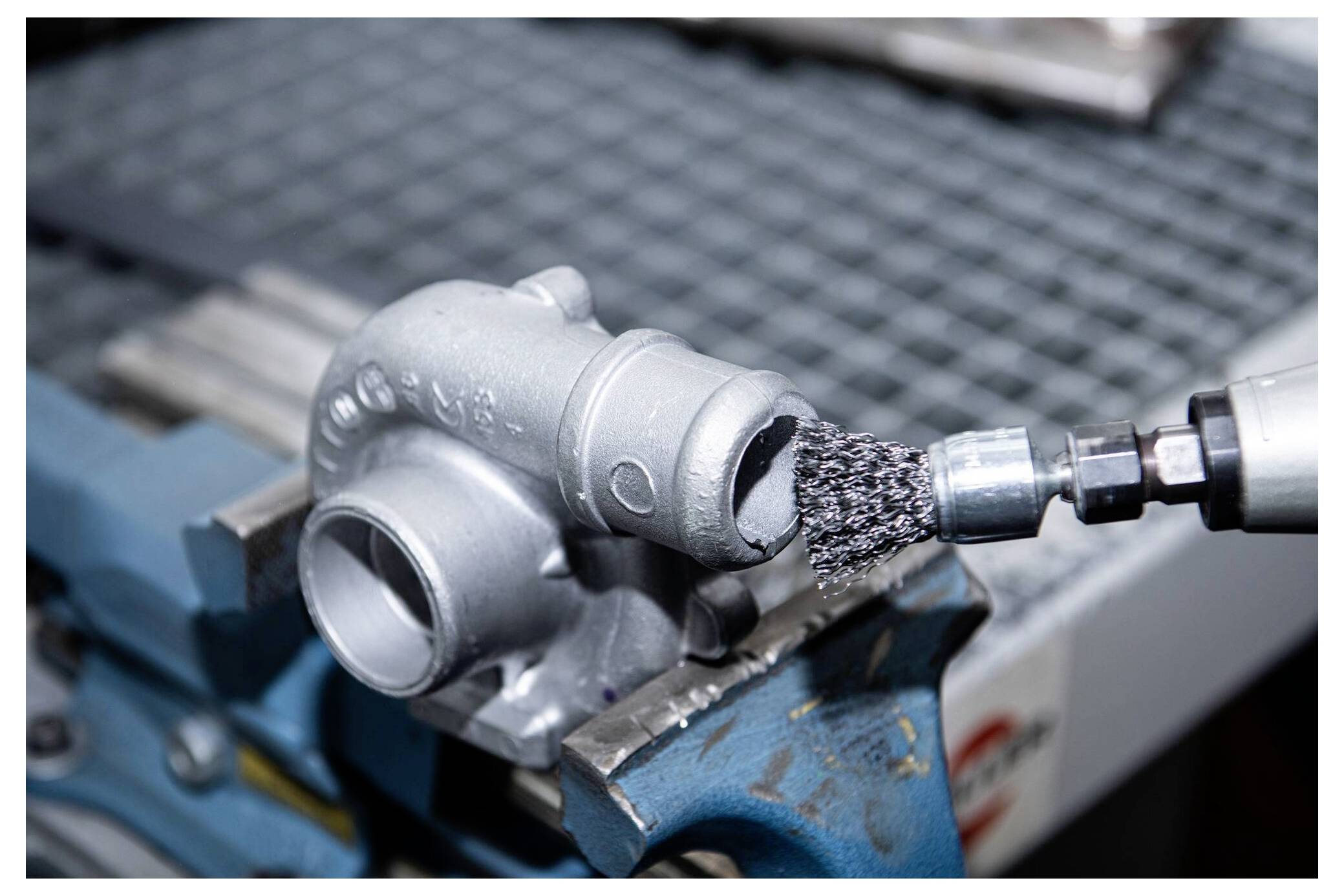 Eine Metallkomponente, die in einem Schraubstock befestigt ist, wird mit einer Drahtbürste gereinigt, die an einem Elektrowerkzeug befestigt ist, vermutlich zur Oberflächenvorbereitung oder -bearbeitung.