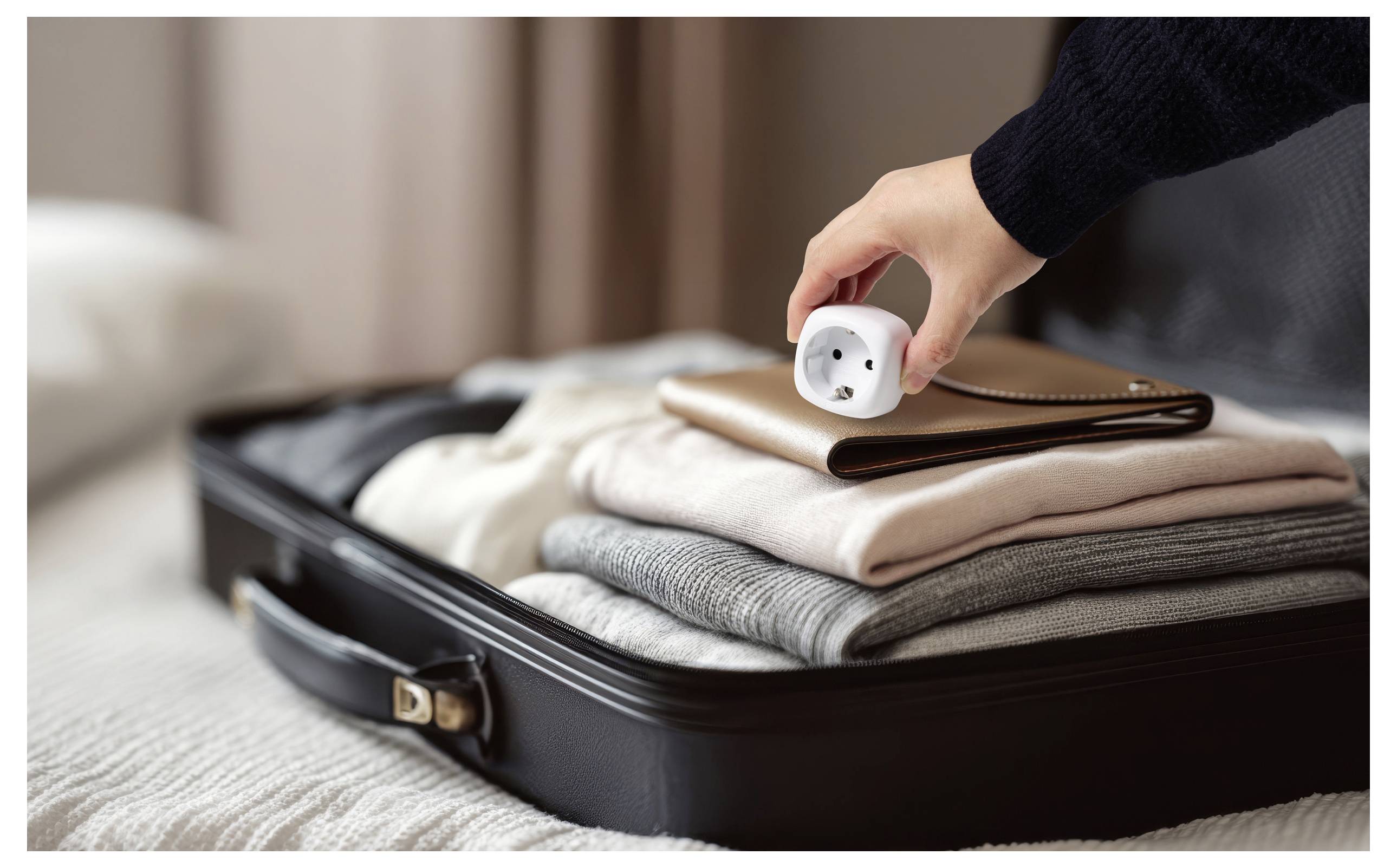 Eine Hand legt einen Reiseadapter auf ordentlich zusammengelegte Kleidungsstücke in einem geöffneten Koffer auf einem Bett, was die Vorbereitung für eine Reise andeutet.