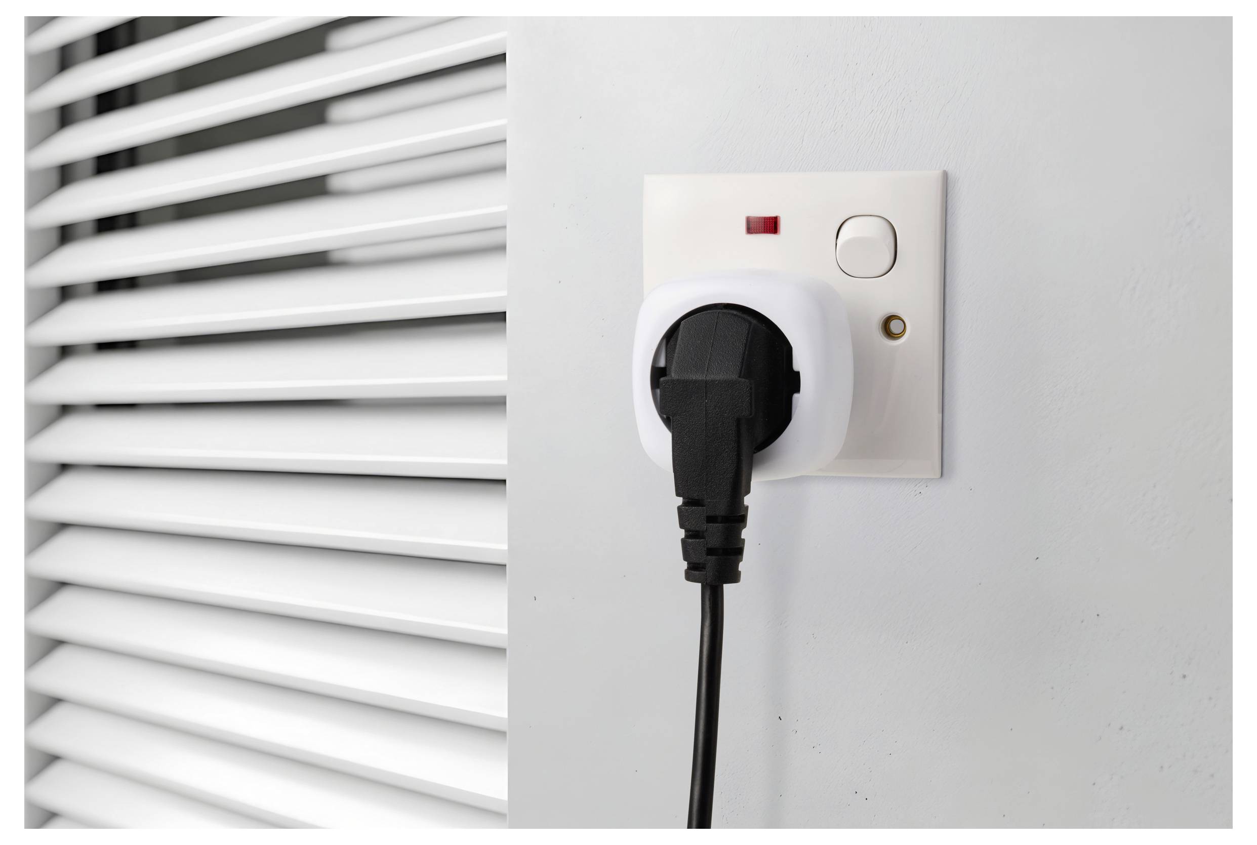 Ein schwarzer Netzstecker wird in eine weiße Wandsteckdose mit roter Kontrollleuchte eingesteckt, neben vertikalen Jalousien an einer grauen Wand.