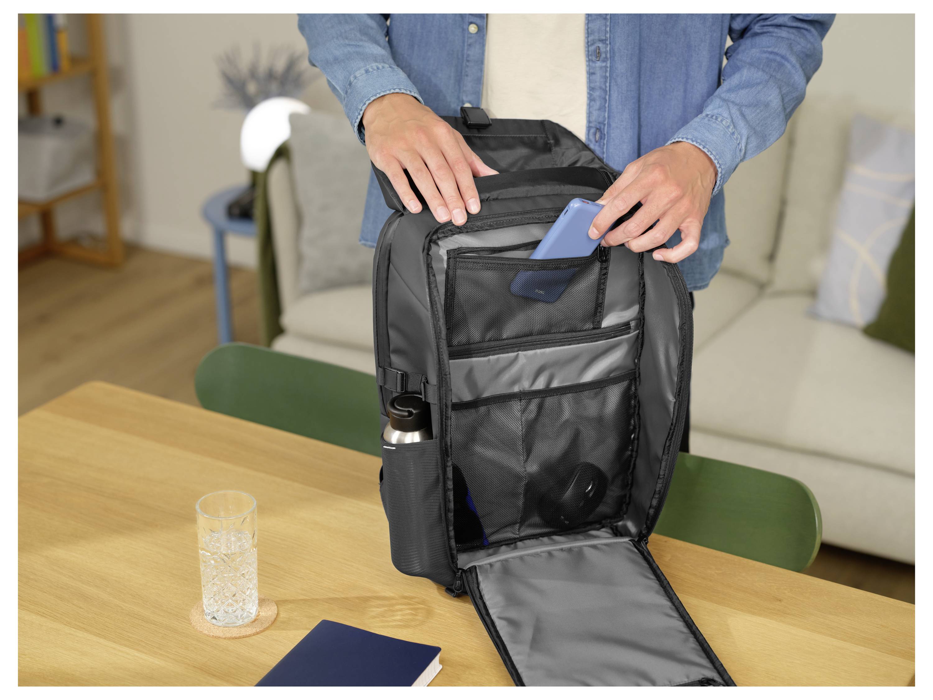 Eine Person steht an einem Tisch und öffnet einen schwarzen Rucksack, um ein blaues Objekt hineinzulegen. Eine Wasserflasche befindet sich in der Seitentasche, und ein Glas sowie ein Notizbuch liegen auf dem Tisch.