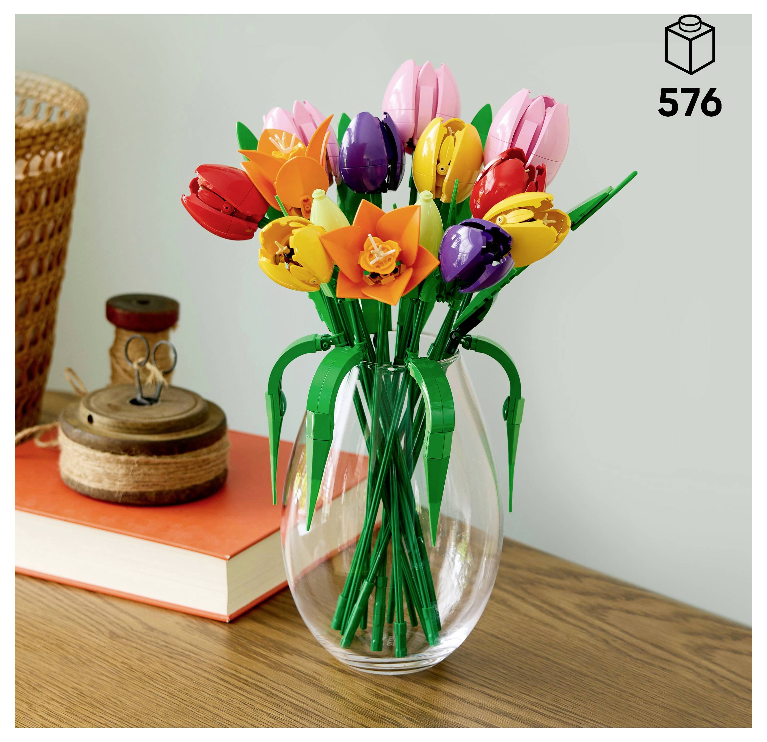 Ein Blumenstrauß aus bunten LEGO-Ziegelblumen in einer klaren Vase, platziert auf einem Holztisch neben einem Buch, einem Flechtkorb und einem Korkbehälter.