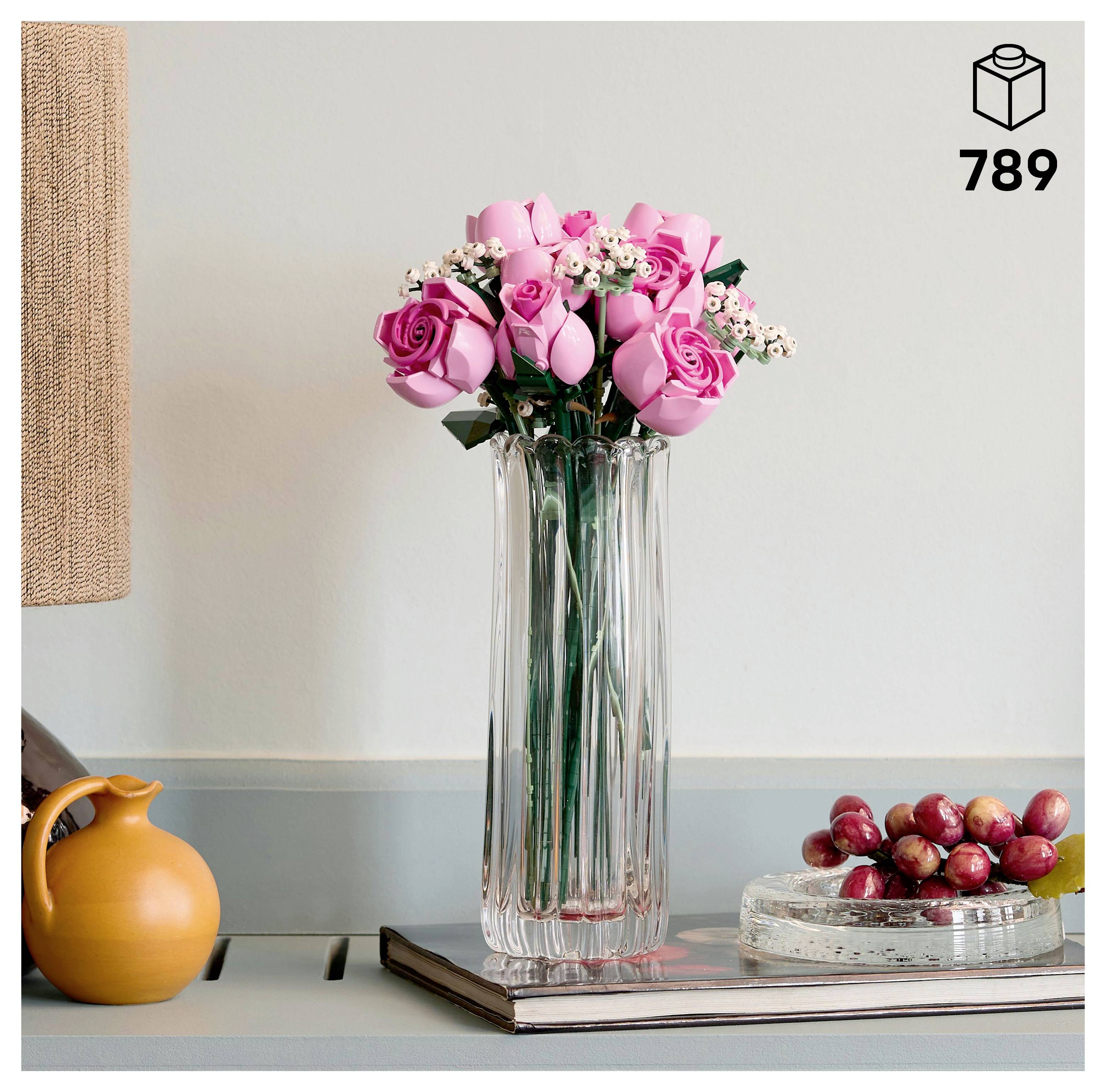 Eine klare Glasvase enthält rosa Rosen und weiße Blumen auf einem Buch. Ein gelber Krug und ein kleiner Teller mit Trauben befinden sich daneben auf einem Regal.