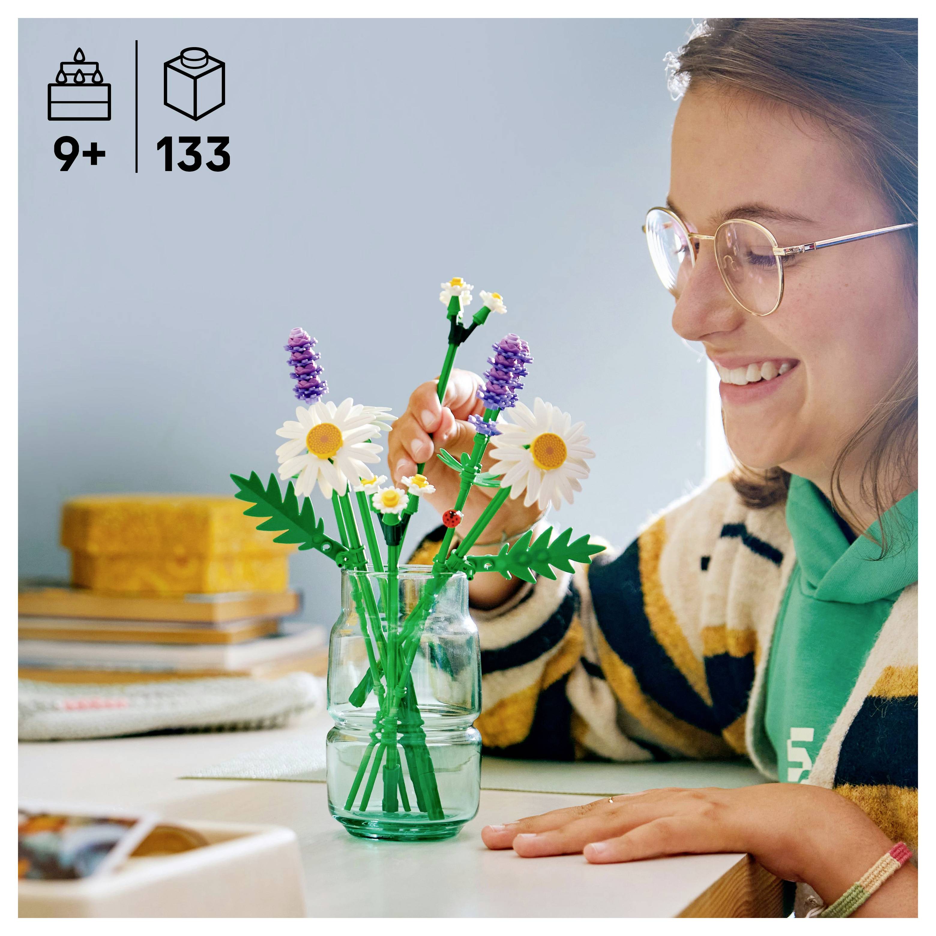 Eine Person, die ein LEGO-Blumenset mit Gänseblümchen und Lavendelblumen zusammenbaut, sitzt an einem Tisch mit einem fröhlichen Gesichtsausdruck.