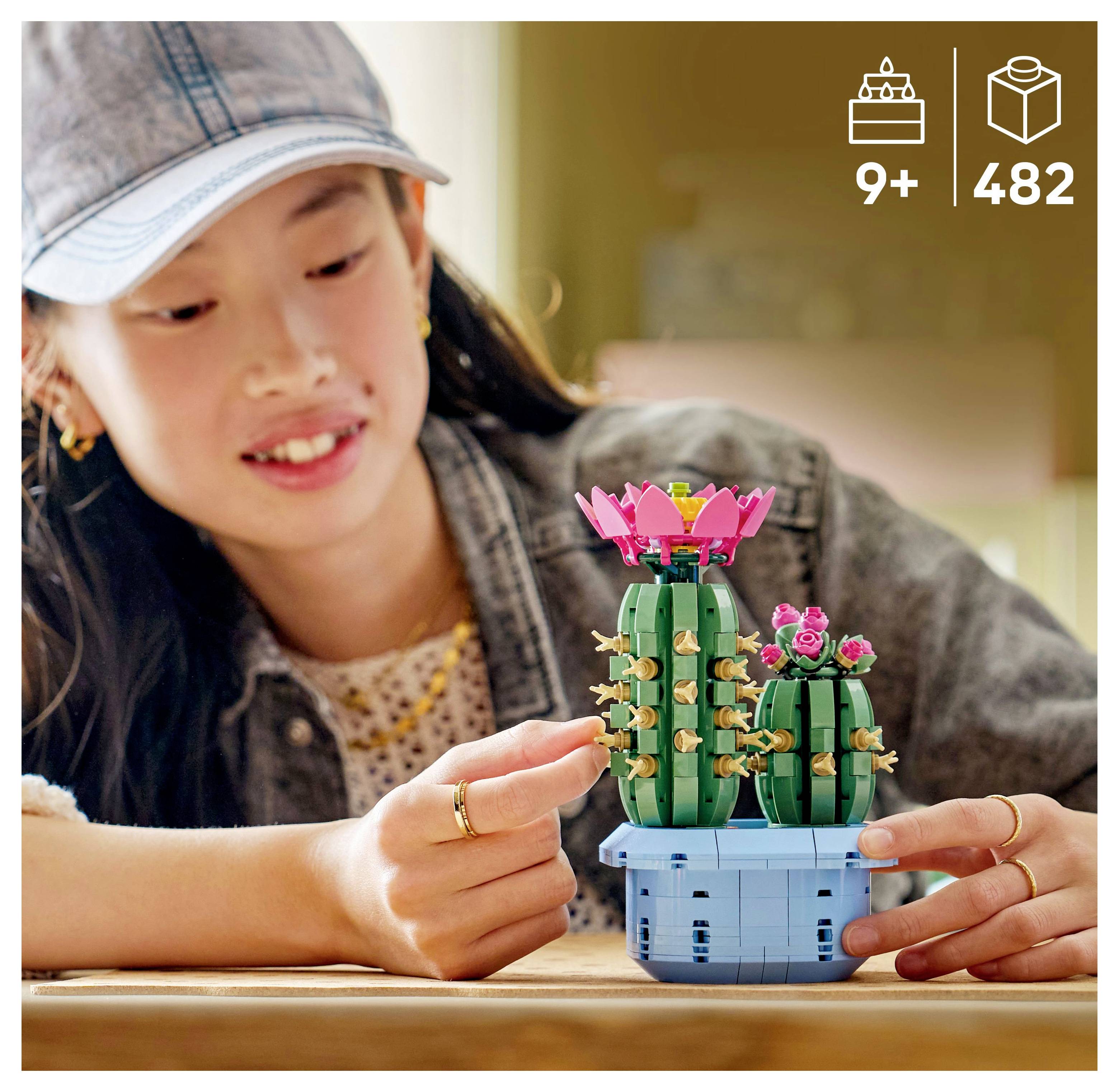 Ein Kind lächelt, während es einen LEGO-Bausatz mit Kakteen und rosa Blüten zusammenbaut. Die Verpackung zeigt die Altersempfehlung „9+