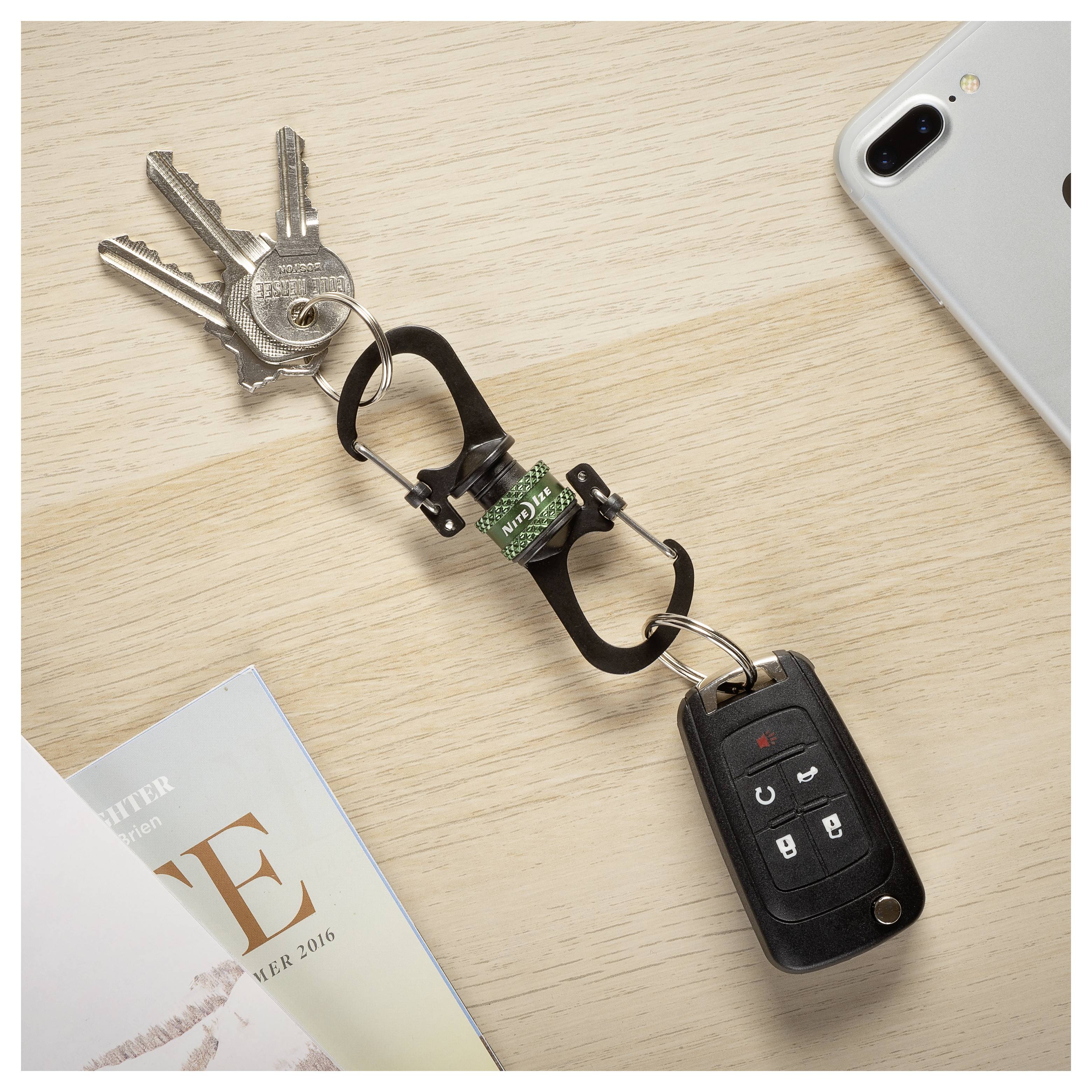 Ein Schlüsselbund an einem schwarzen Karabiner und ein Autoschlüssel liegen auf einer hellen Holzoberfläche, in der Nähe einer Zeitschrift und eines Smartphones.