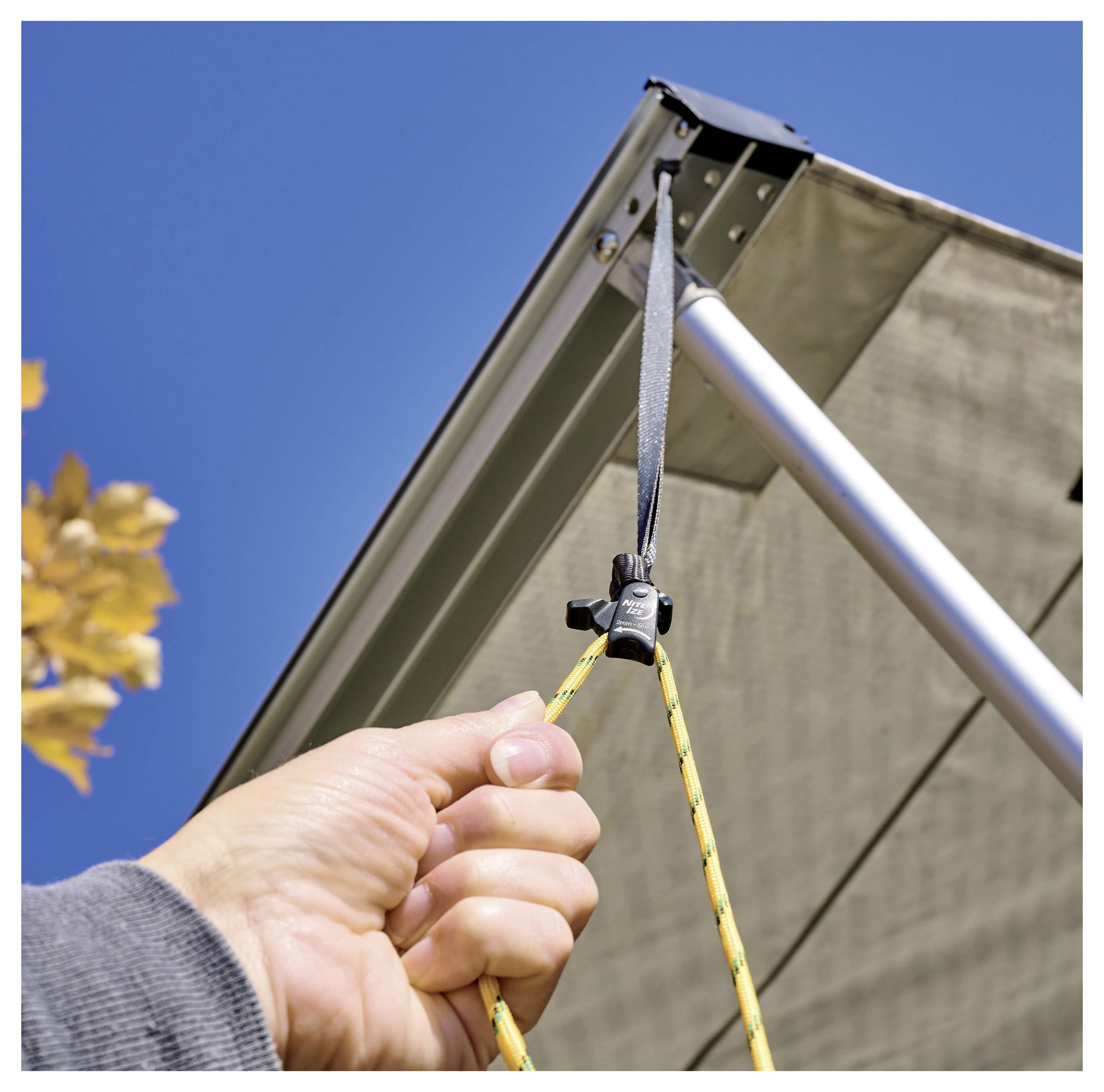 Eine Hand justiert die Spannung an einem Seil, das an einem Sonnensegel befestigt ist, mit einem klaren blauen Himmel und einem Hauch von Blattwerk im Hintergrund.