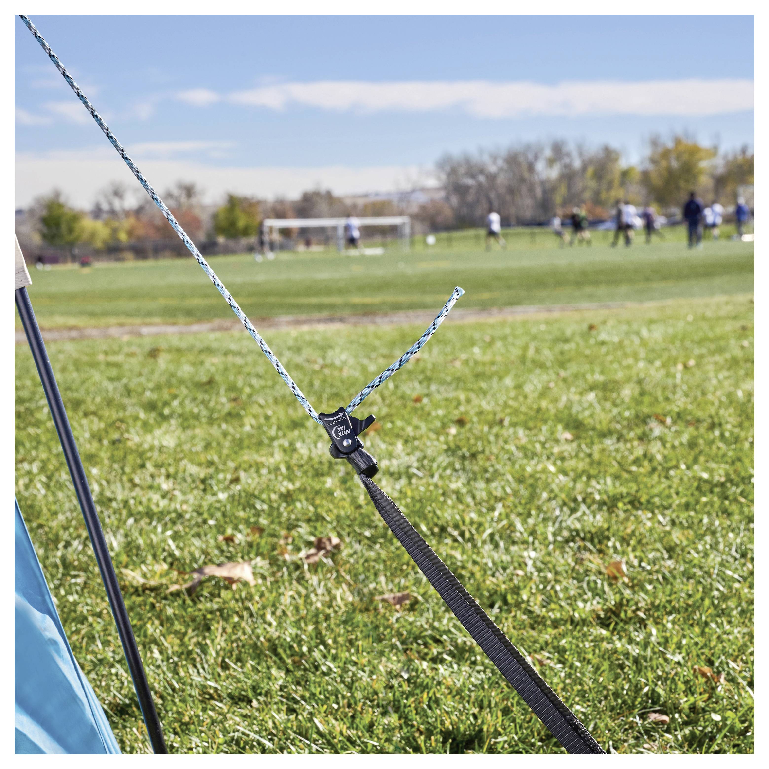 Ein blau-blaues Zelt, das auf einem grasbewachsenen Feld aufgestellt ist, mit einem Seil, das durch einen Spanner gesichert wird. Fußballtore und Spieler sind im Hintergrund zu sehen.