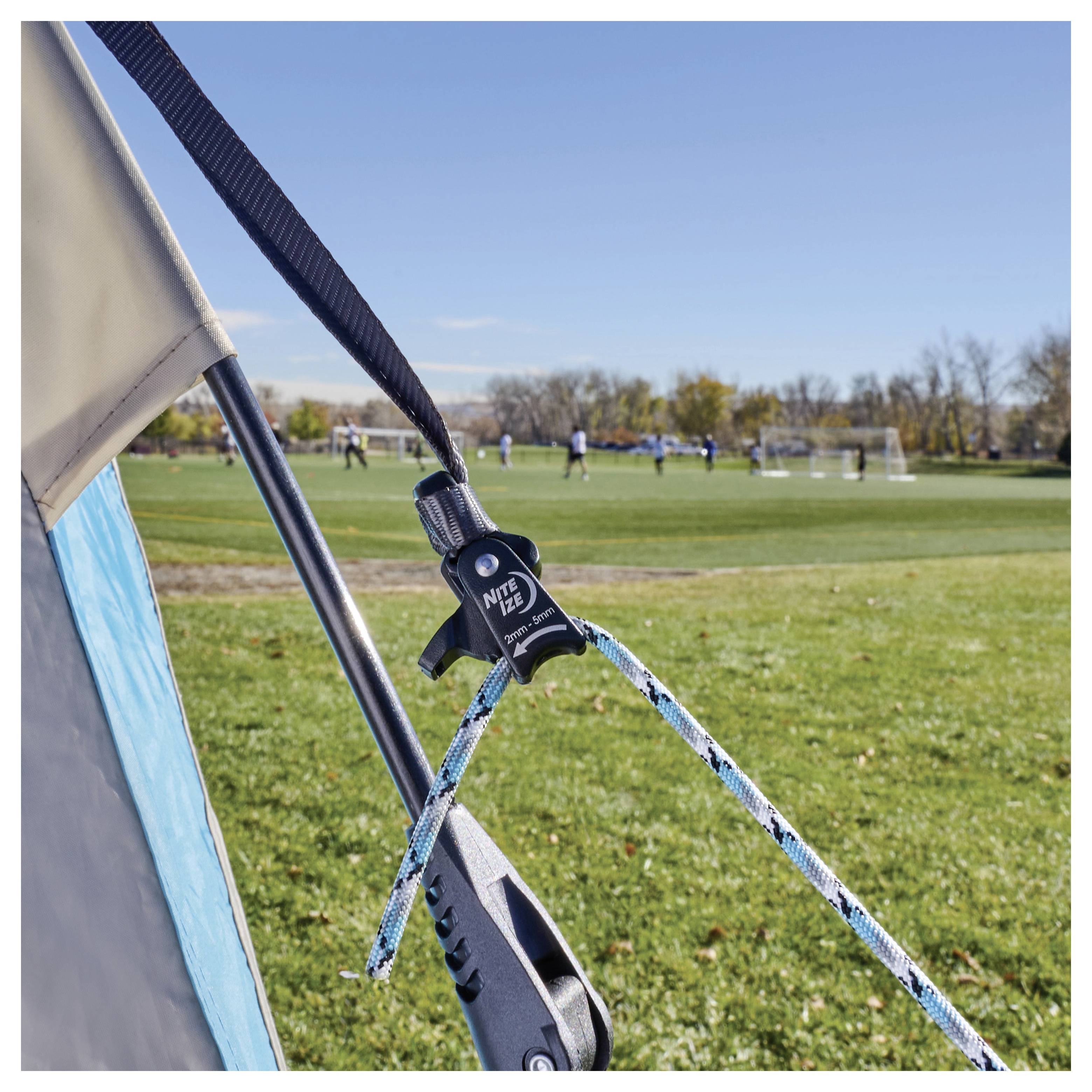 Nahaufnahme eines Herds- und Seils, das ein Zelt auf einem Rasenfeld sichert, wobei ein Fußballspiel im Hintergrund zu sehen ist.