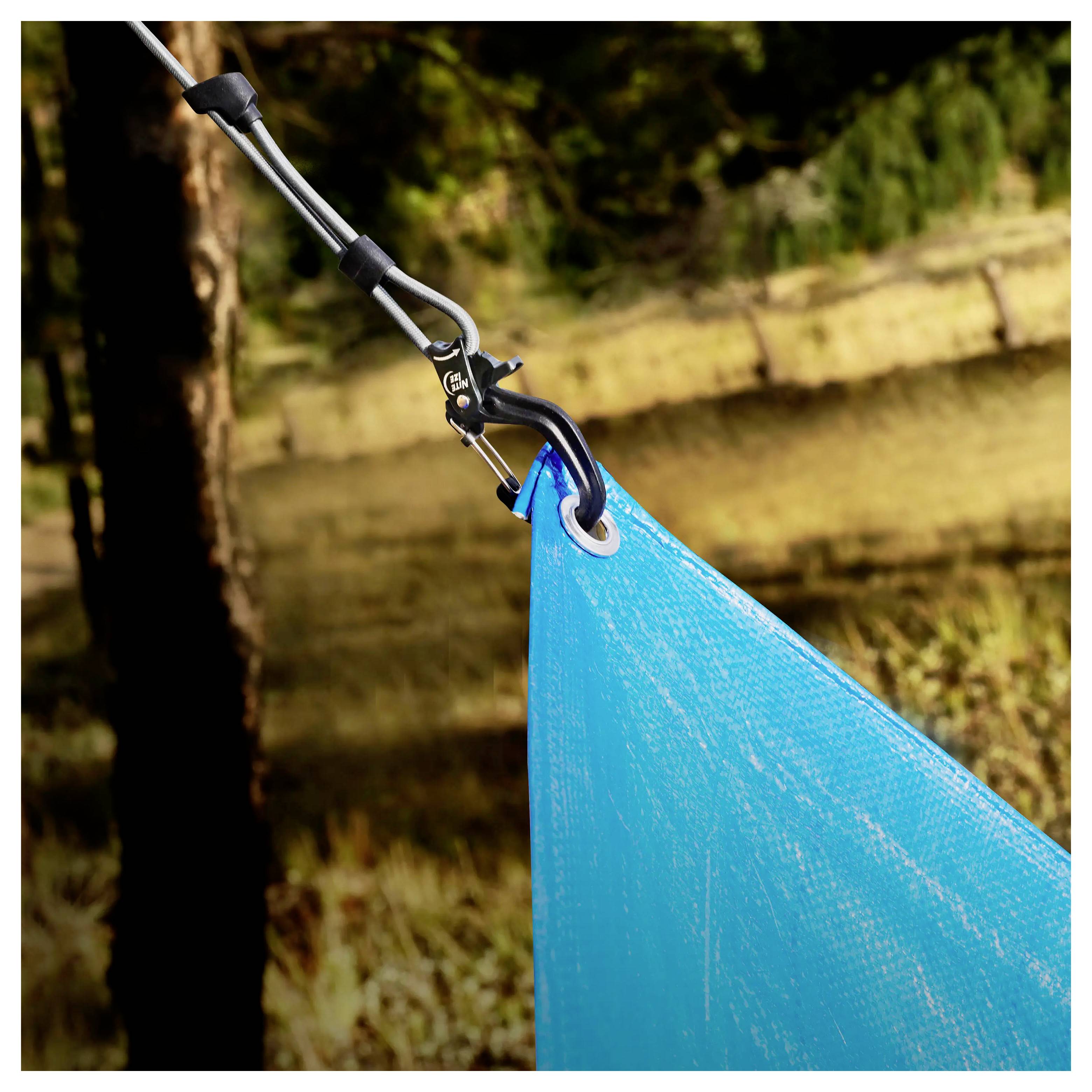 Eine Nahaufnahme einer blauen Plane, die mit einem Karabiner an einem Seil befestigt ist, welches an einem Baum im Freien angebunden ist, mit einem unscharfen Waldhindergrund.