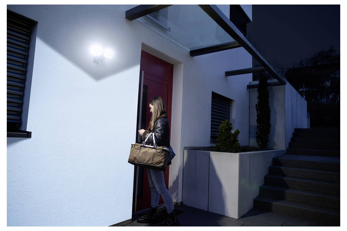 Eine Frau mit einer Tasche betritt bei Nacht ein modernes Haus durch eine rote Tür, beleuchtet von einem hellen, bewegungsaktiviertem Sicherheitslicht.