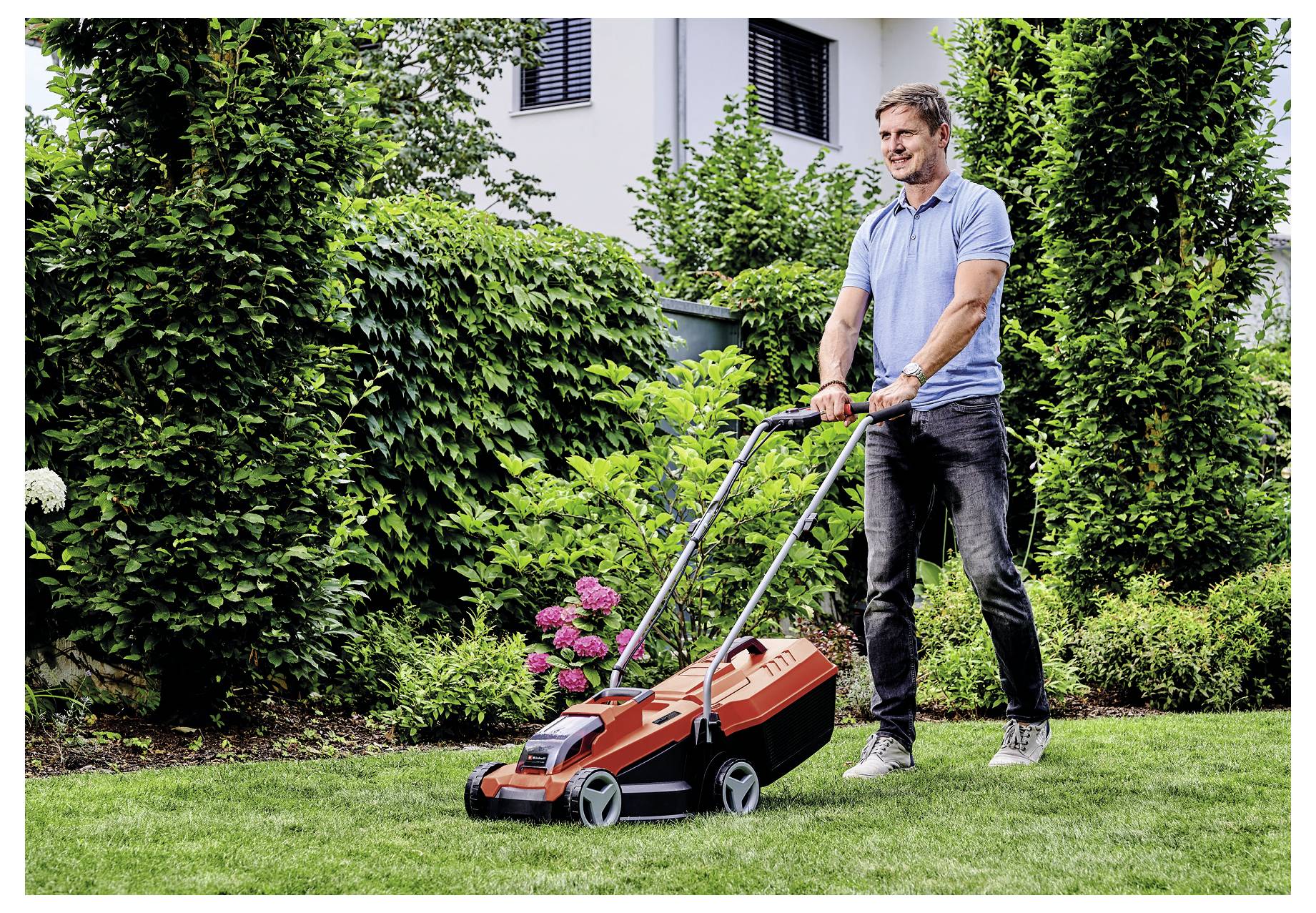 Ein Mann schiebt einen Rasenmäher auf einem grasbewachsenen Rasen. Er ist im Freien, umgeben von Grünflächen und einem blühenden Strauch, mit einem Haus im Hintergrund.