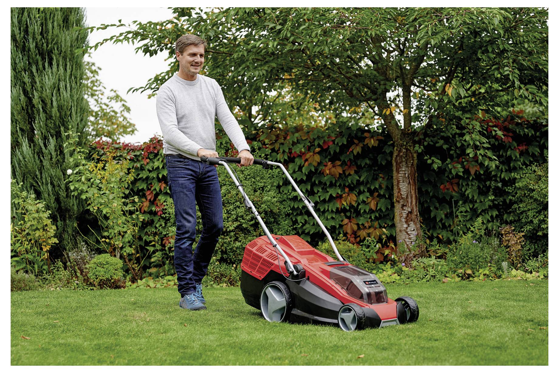 Mann, der einen Rasen mit einem roten elektrischen Rasenmäher mäht, umgeben von grünem Gras, Bäumen und einer Hecke.