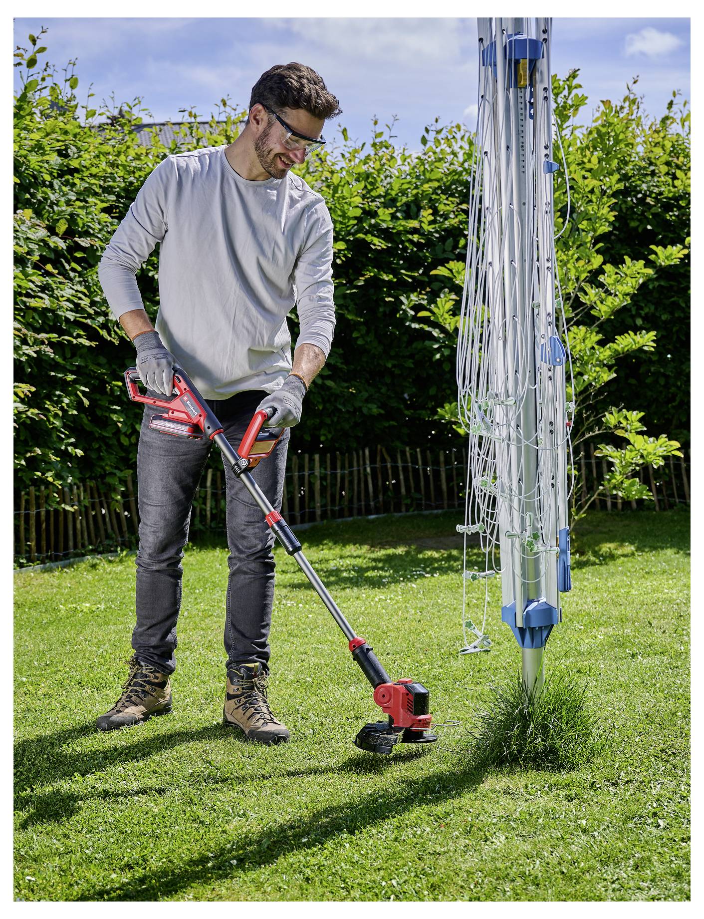 Eine Person mit Schutzhandschuhen und Schutzbrille verwendet einen roten elektrischen Rasentrimmer, um Gras in der Nähe einer Wäscheleine in einem sonnigen Garten zu schneiden.