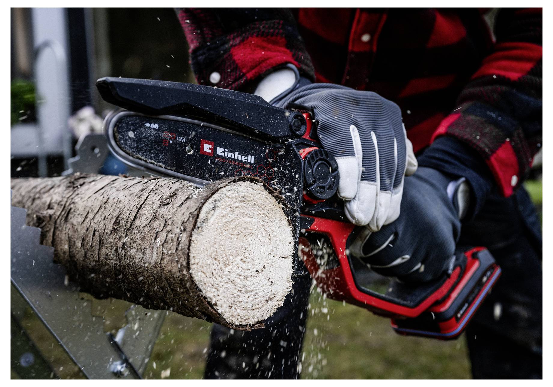 Eine Person mit Schutzhandschuhen sägt einen Baumstamm mit einer kleinen Kettensäge im Freien. Sägespäne fliegen, während die Kettensäge durch das Holz schneidet.