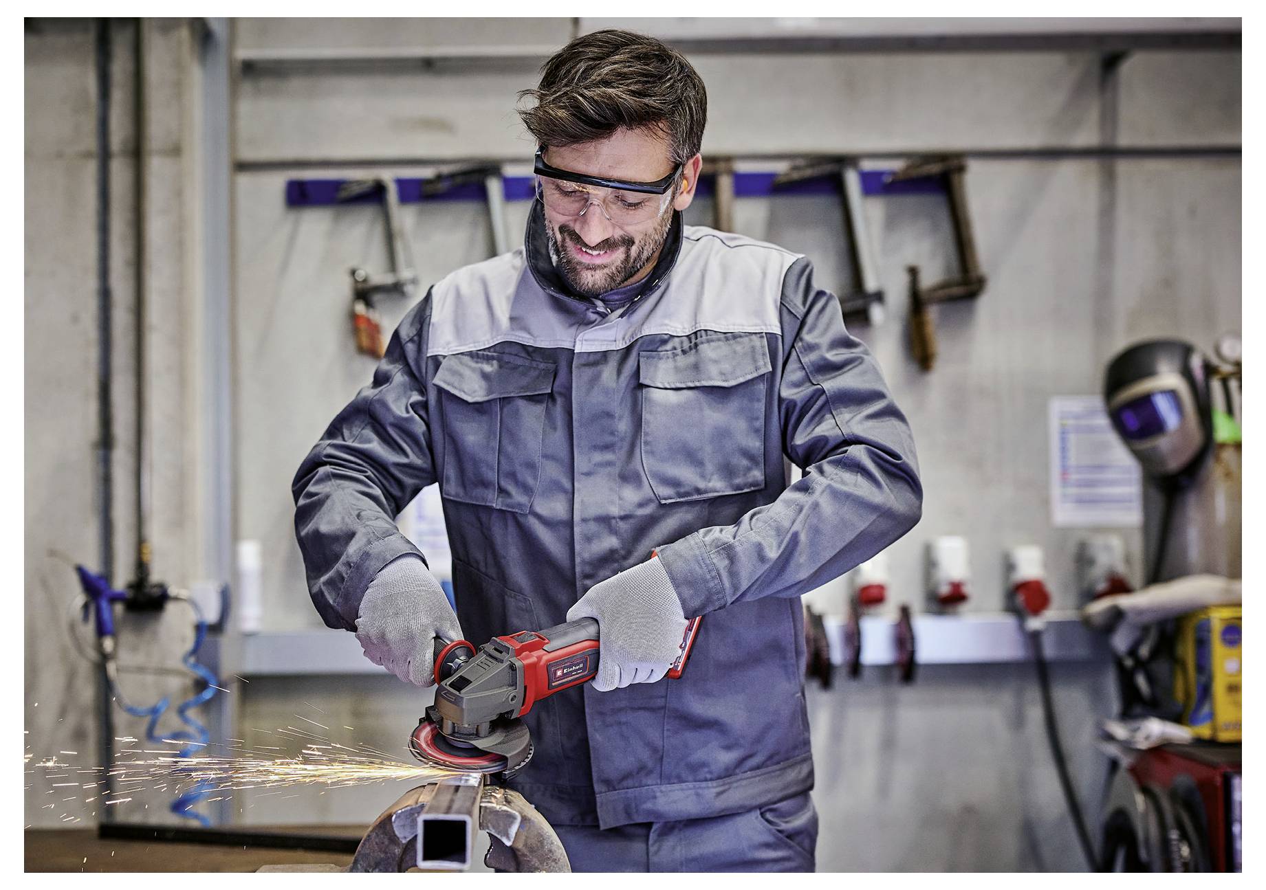 Ein Arbeiter mit Schutzbrille und Handschuhen benutzt einen Winkelschleifer an einem Metallstück in einer Werkstatt. Funken sind während des Schleifvorgangs sichtbar.
