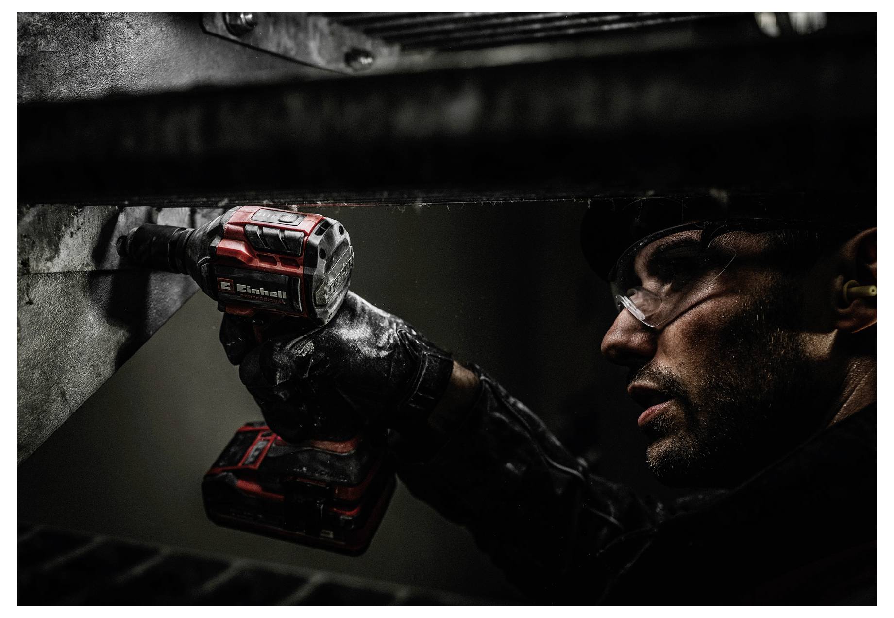 Eine Person verwendet einen roten drahtlosen Bohrer auf einer Metalloberfläche, trägt Schutzbrille und Handschuhe und ist konzentriert bei einer Aufgabe in einer dunkel beleuchteten Umgebung.