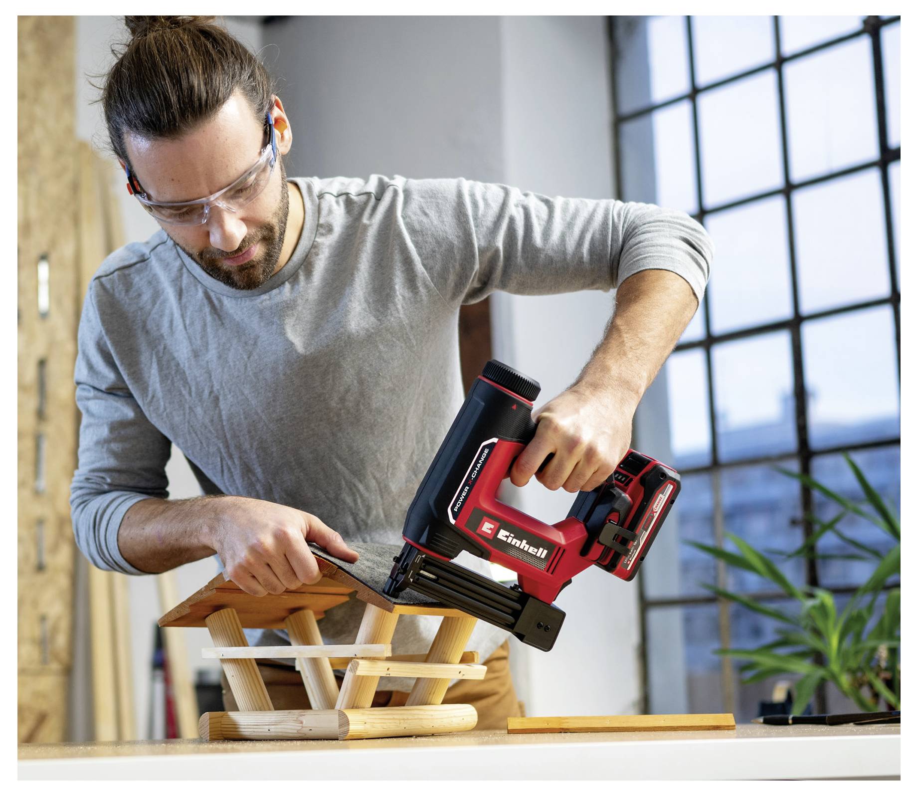 Eine Person mit Schutzbrille benutzt ein Elektrowerkzeug, um drinnen eine Holzkonstruktion zu montieren, wobei ein Fenster und eine Pflanze im Hintergrund zu sehen sind.