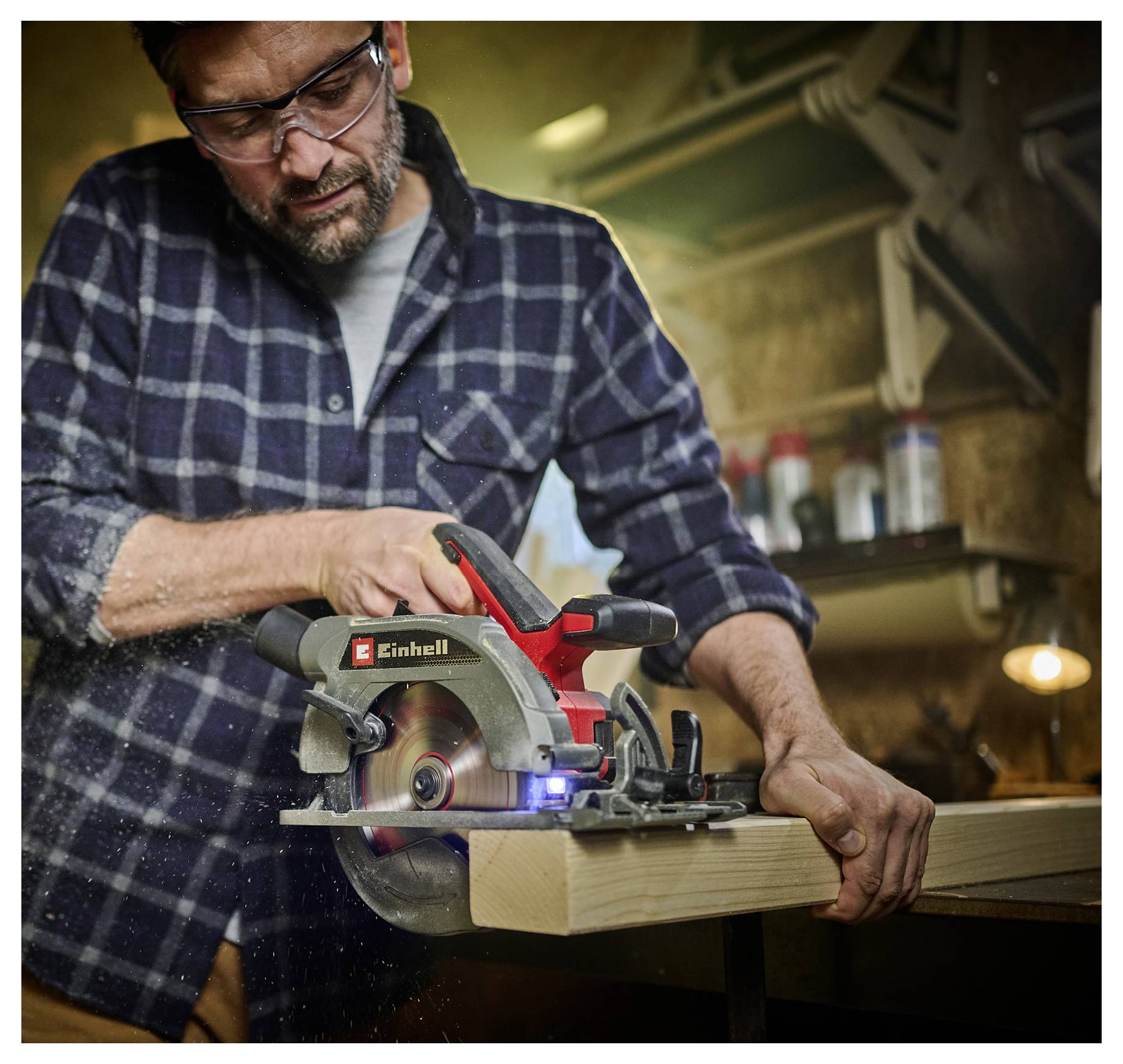 Eine Person mit Sicherheitsschutzbrille und kariertem Hemd verwendet eine Kreissäge, um ein Holzbrett in einer Werkstattumgebung zu schneiden.