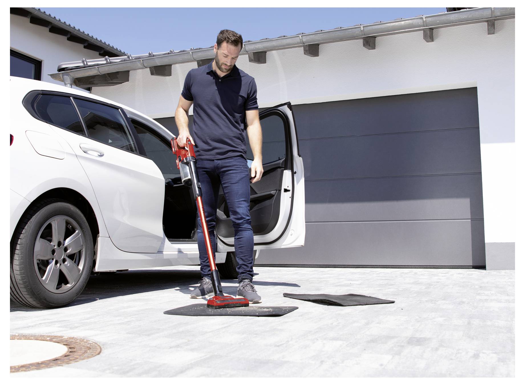 Ein Mann saugt Fußmatten eines Autos im Freien neben einem weißen Auto, das vor einer Garage geparkt ist.