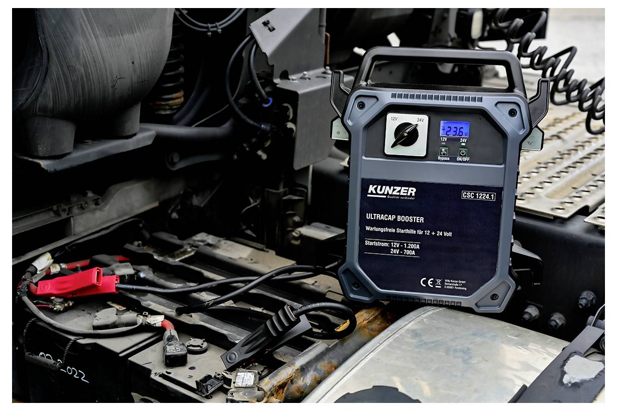 Ein Batteriebooster mit der Bezeichnung 'KUNZER Ultracap Booster' ist an eine Fahrzeugbatterie angeschlossen und zeigt eine Spannung von 12,96 V.