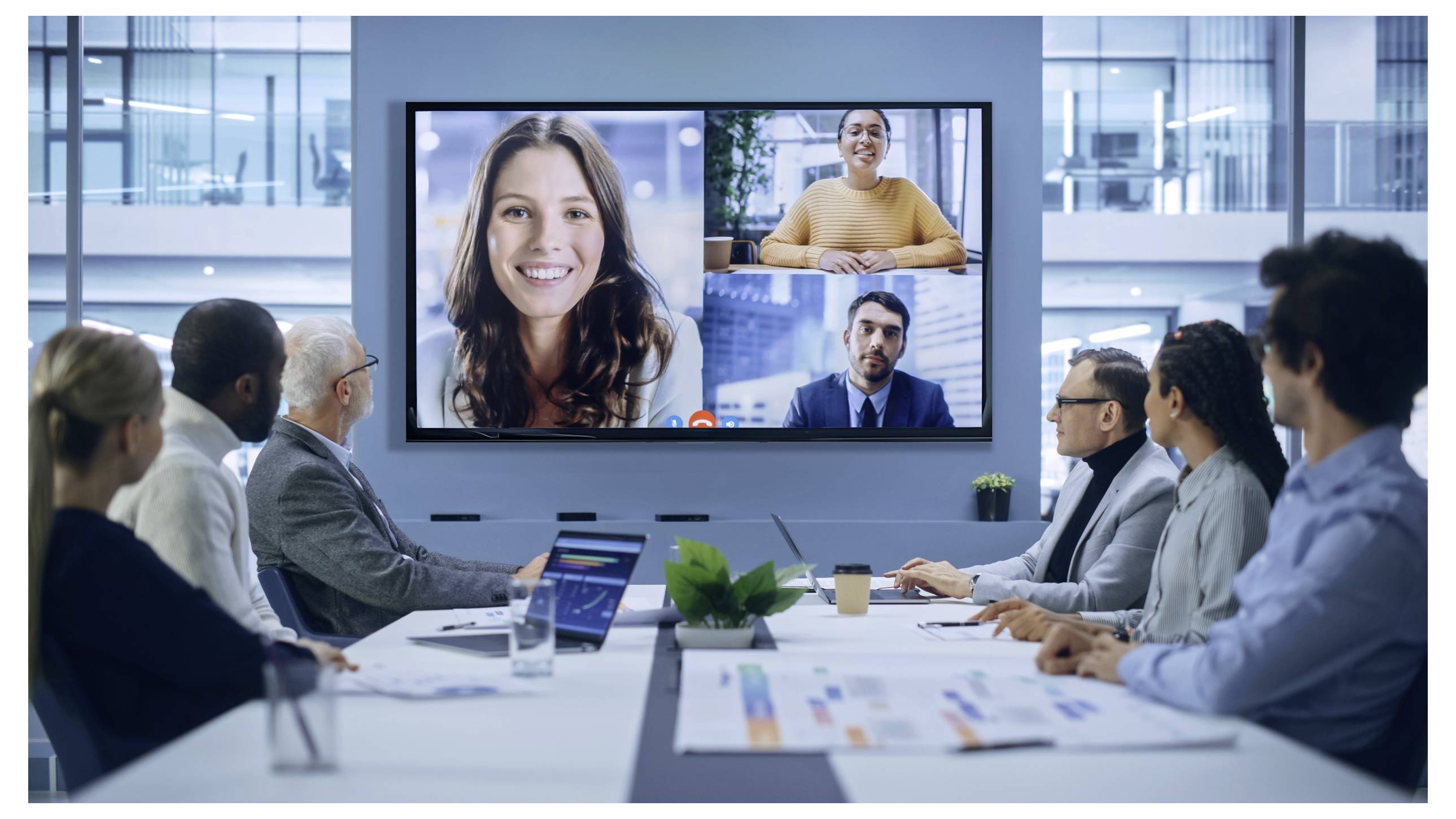 Personen in einem Konferenzraum, die an einem Videoanruf teilnehmen, der auf einem großen Bildschirm angezeigt wird, mit zwei Frauen und einem Mann.