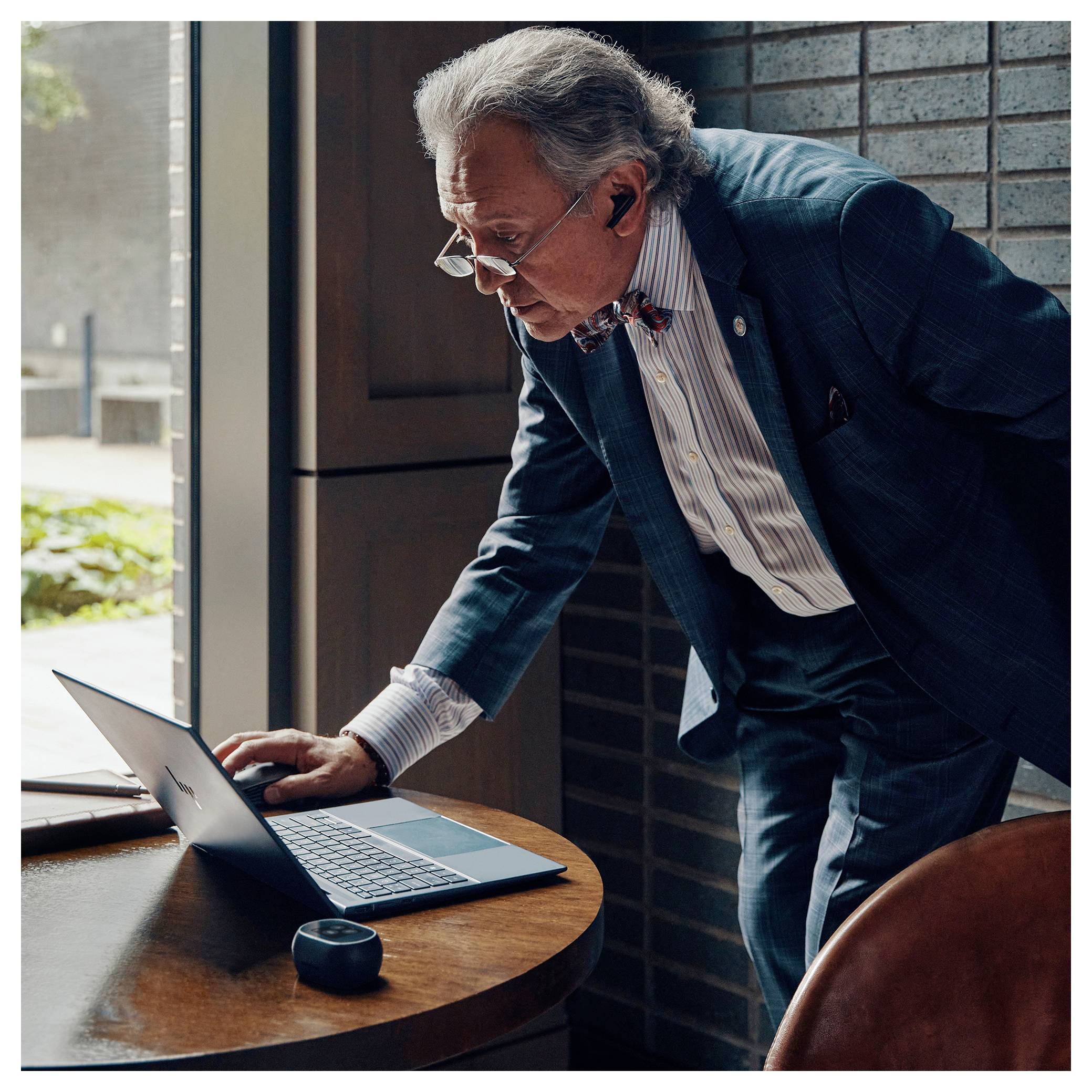 Eine ältere Person in formeller Kleidung lehnt sich über einen Laptop auf einem Holztisch und wirkt konzentriert, mit einem Ausblick auf Grünflächen hinter ihr.