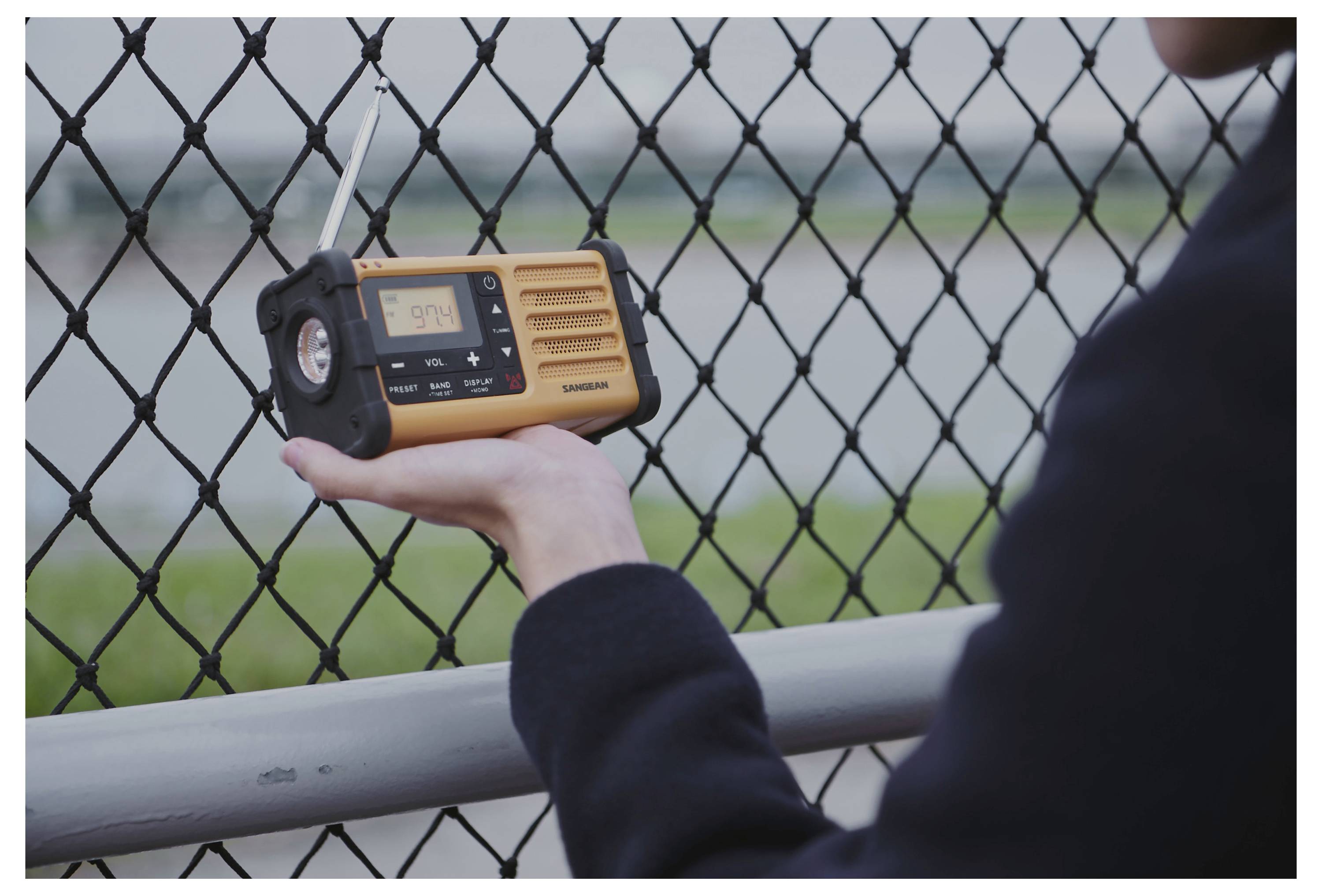 Eine Person hält ein gelbes Notfallradio mit einem Display, auf dem '5:14' angezeigt wird. Im Hintergrund befindet sich ein Zaun und eine unscharf werdende Landschaft.