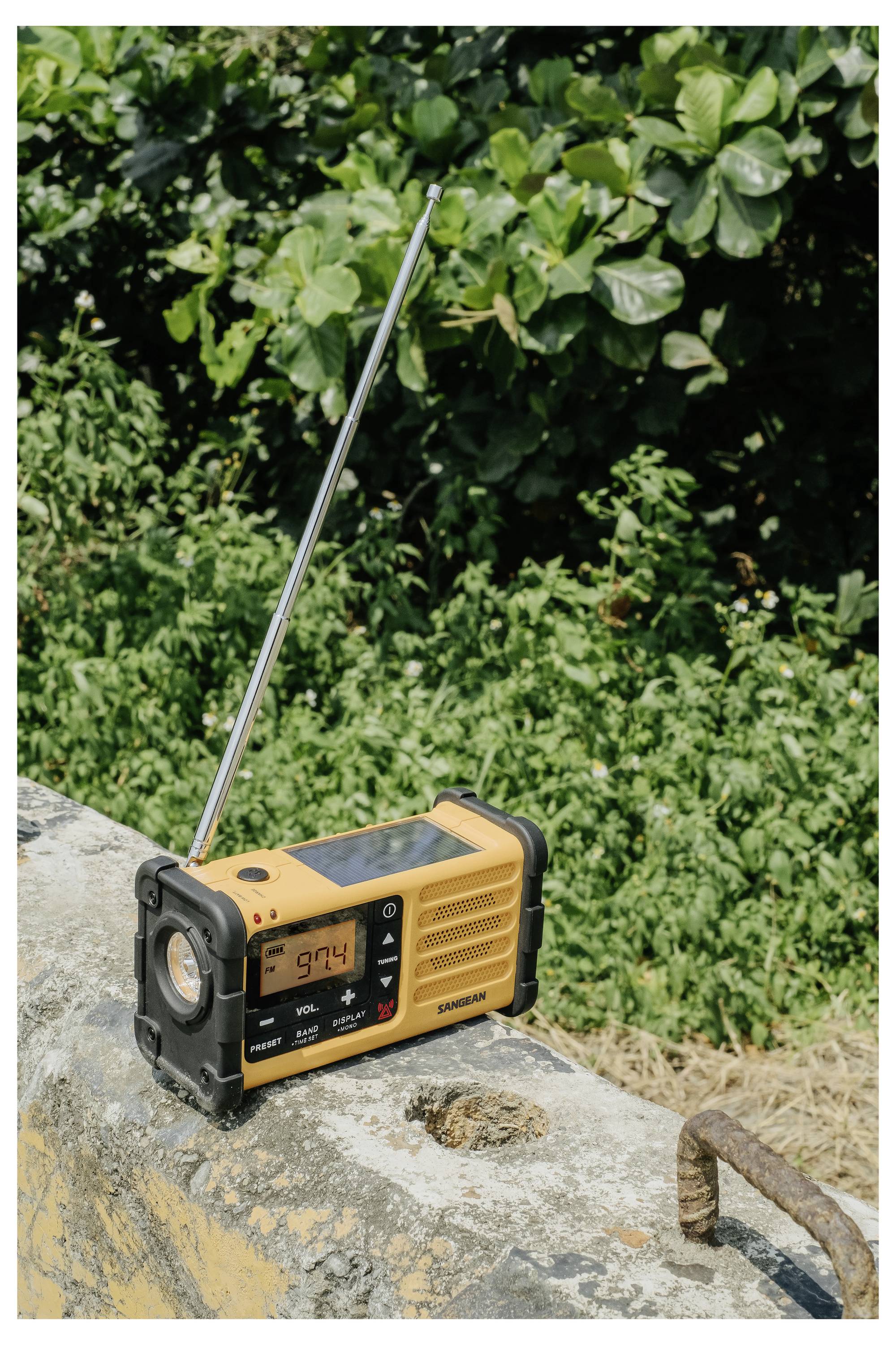 Ein gelbes Radio mit ausgefahrener Antenne steht auf einem Betonvorsprung im Freien, umgeben von Grünpflanzen. Das digitale Display zeigt '97.4'.