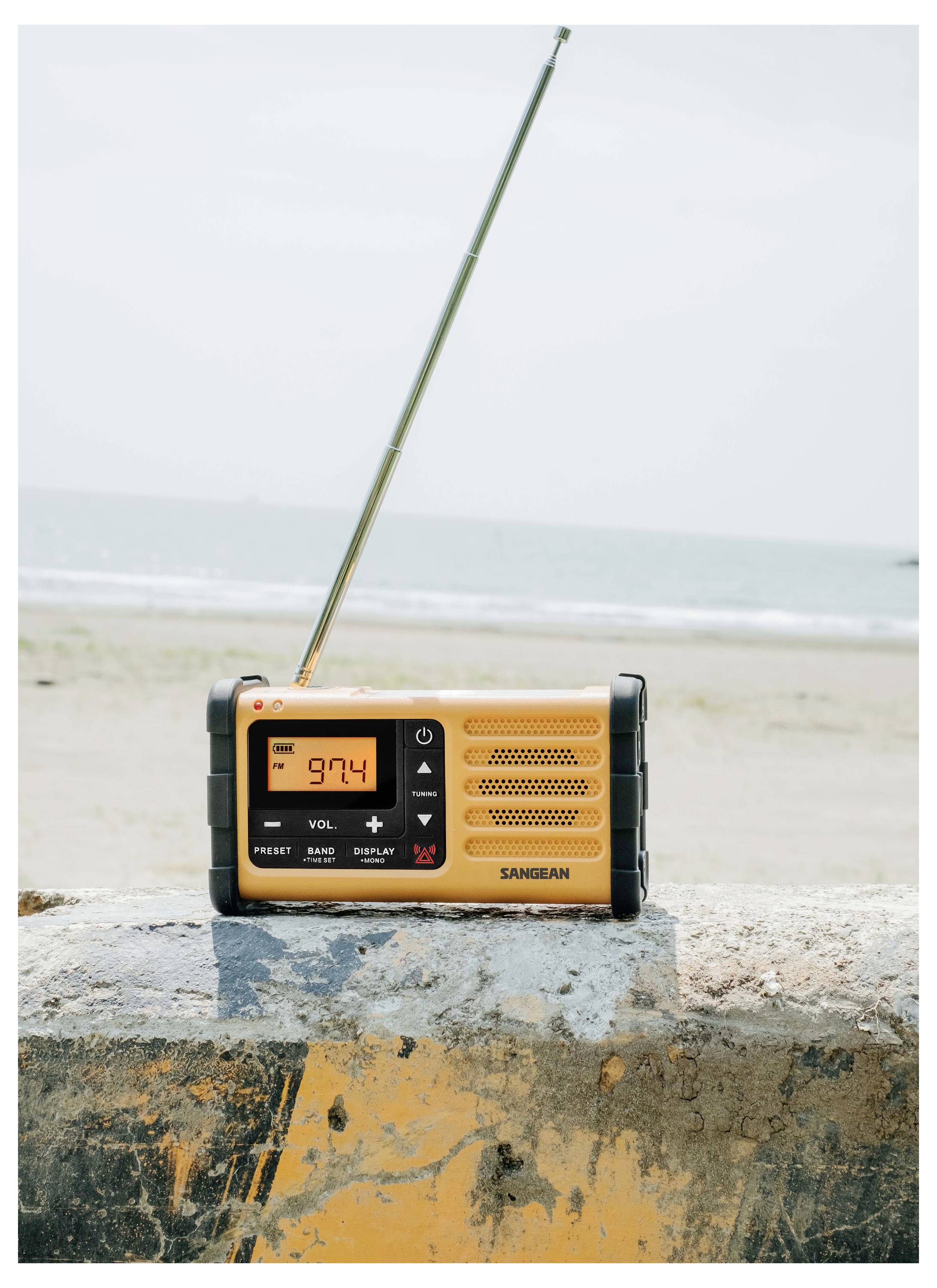 Ein gelbes tragbares Radio mit ausgefahrener Antenne steht auf einer Betonbarriere am Strand. Das Display zeigt '97.4'.