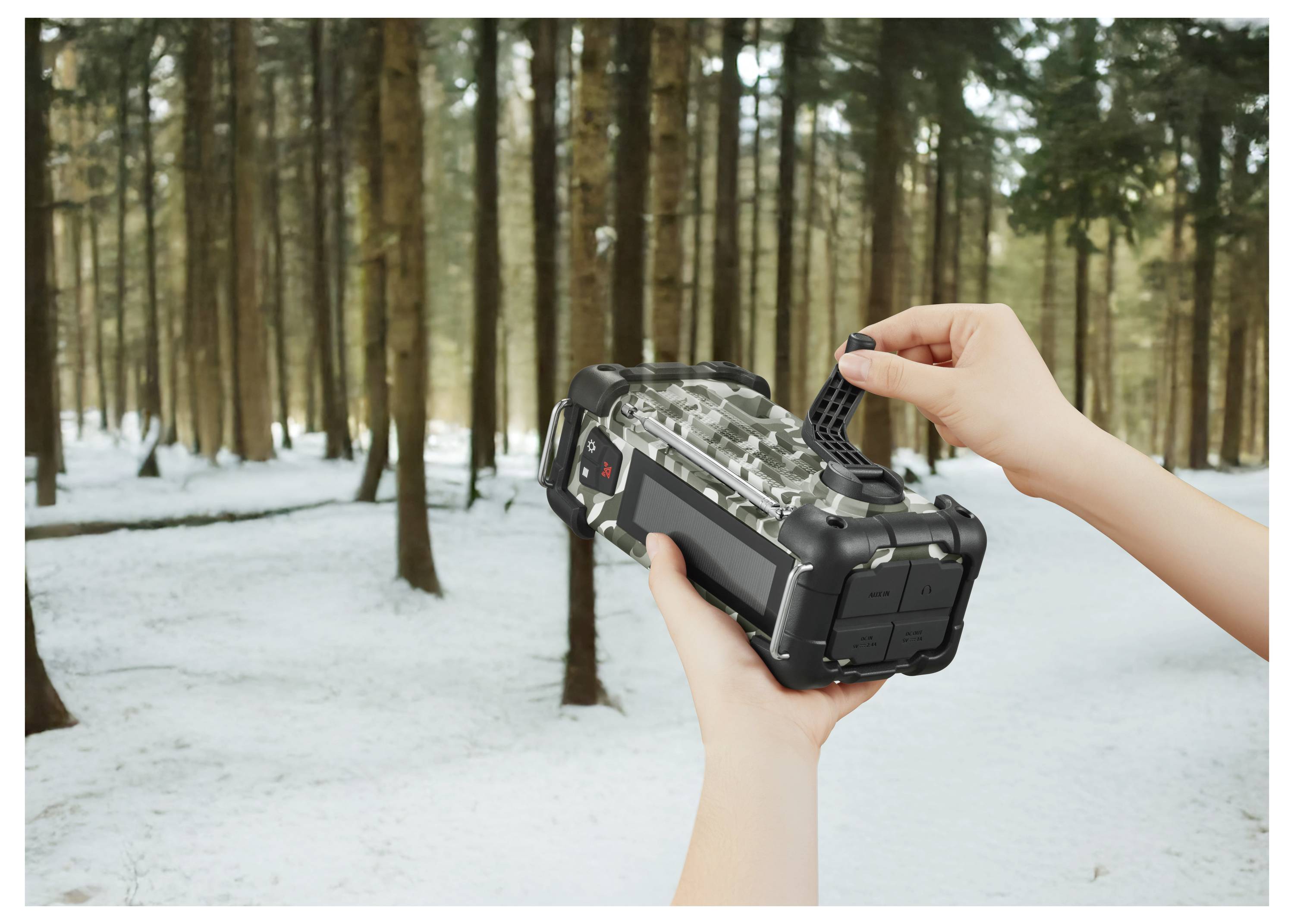 Eine Person hält eine schwarze Wildkamera in einem schneebedeckten Wald und justiert sie. Im Hintergrund sind hohe Bäume zu sehen.