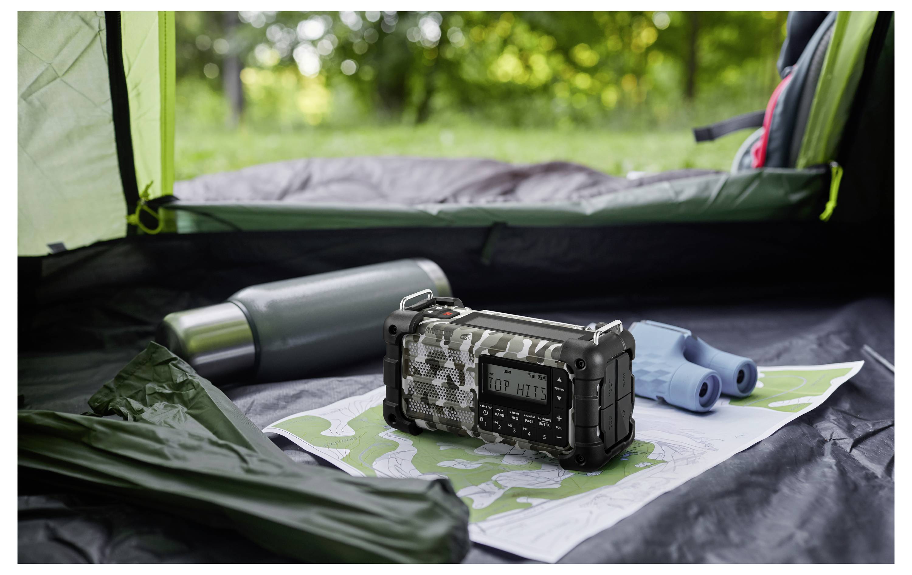 Ein tragbares Radio mit Tarnmuster sitzt auf einer Karte in einem Zelt, begleitet von einem Thermoskanne und einem Fernglas; üppiges Grün ist draußen zu sehen.