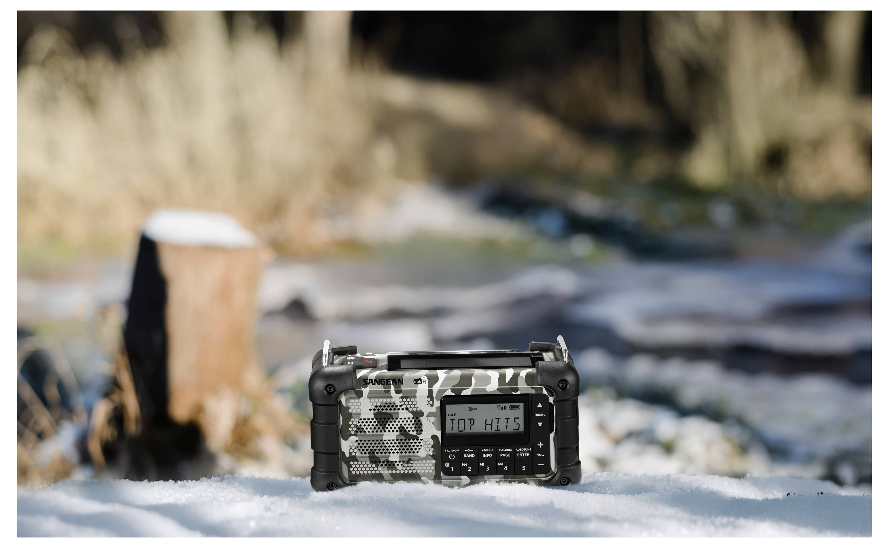 Ein tragbares Radio mit Tarndesign liegt auf Schnee, mit einem unscharfen natürlichen Hintergrund mit Bäumen und einem Baumstumpf.