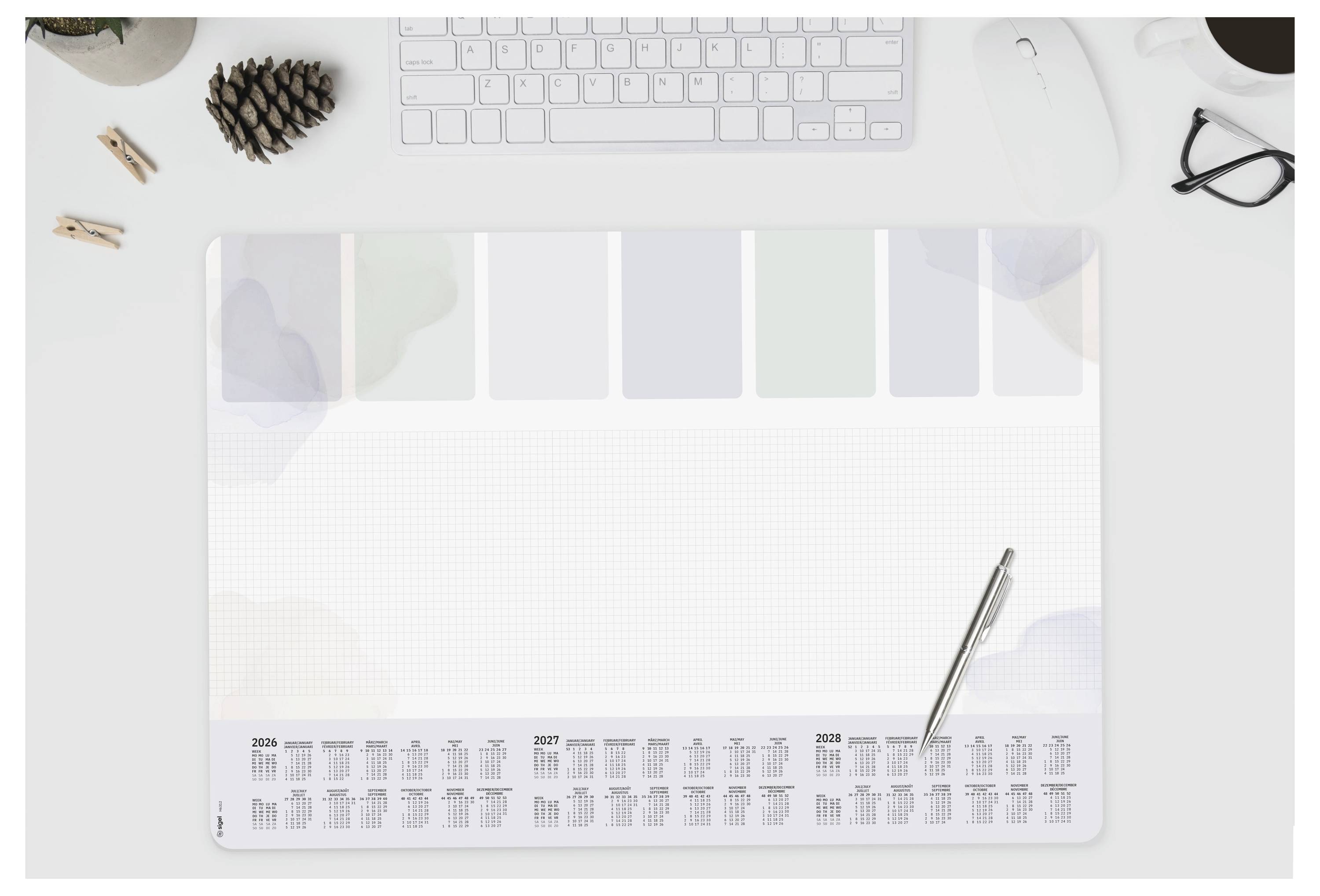 Ein Kalender-Schreibtischunterlage mit einem Stift darauf, eine Computertastatur, Computermaus, Tannenzapfen, Brille und ein Paar Clips auf einem weißen Schreibtisch.