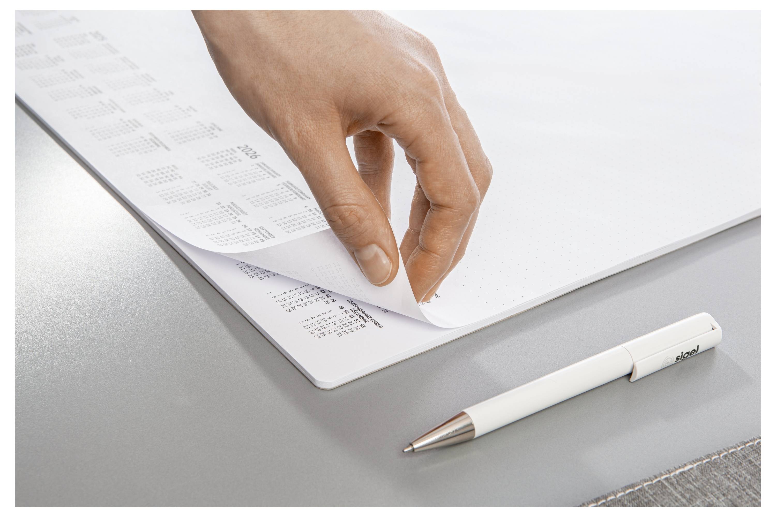 Eine Hand schlägt eine Seite eines Kalenders auf einem Schreibtisch um, neben einem weißen Stift.