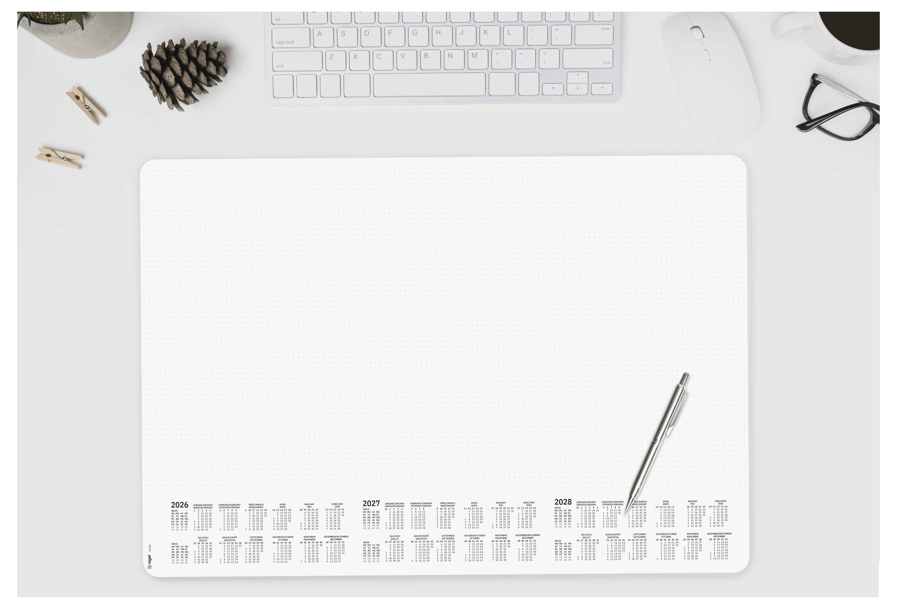 Ein Schreibtischorganizer verfügt über einen Kalender von 2023 bis 2025, einen Stift rechts daneben und einen Tastatur, Maus, Kaffeetasse und Tannenzapfen im Sichtfeld.