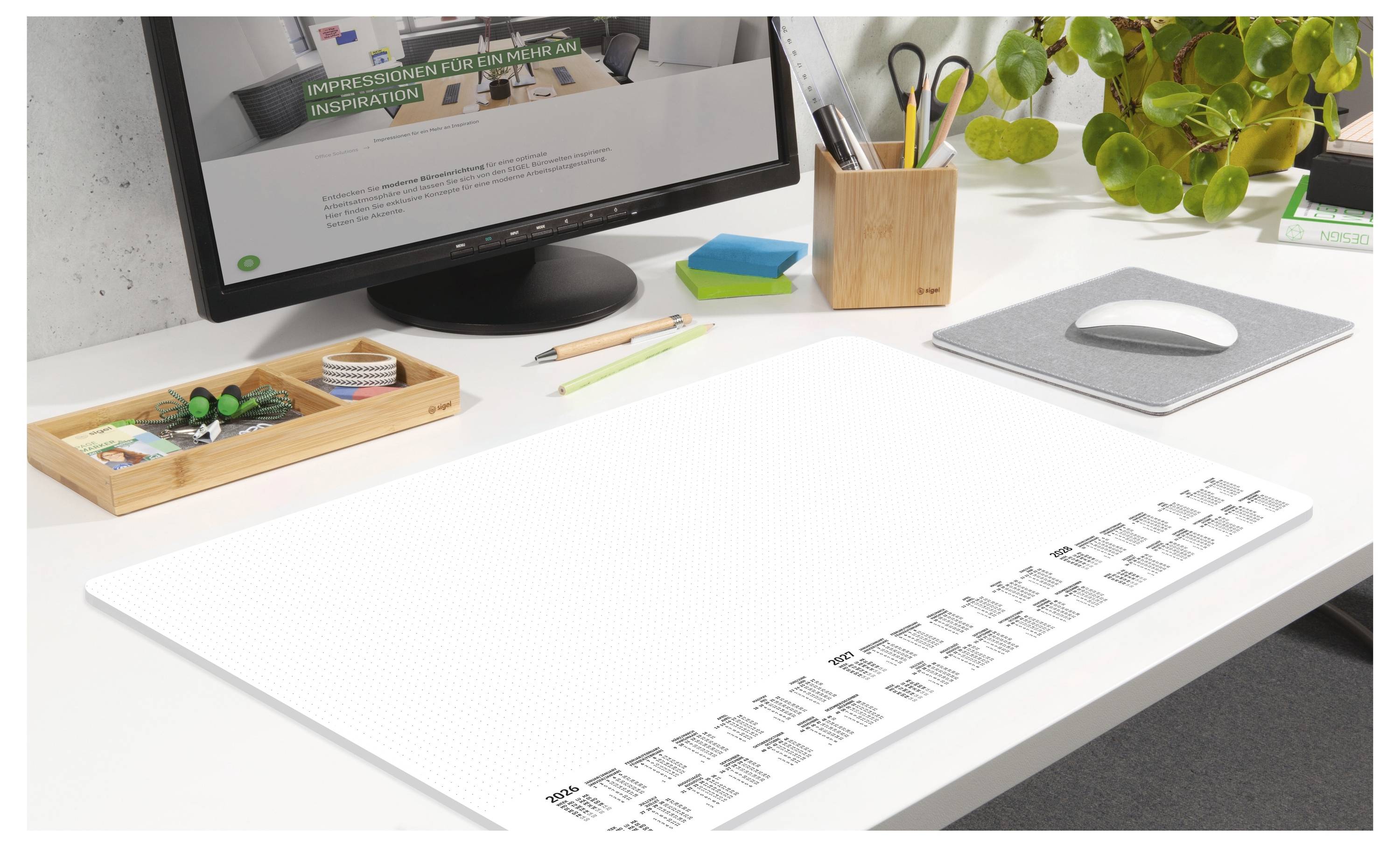 Ein modernes Schreibtisch-Setup mit einem Computermonitor, einer Maus und ordentlich arrangiertem Schreibmaterial. Eine Pflanze verleiht dem Ganzen einen Hauch von Grün.