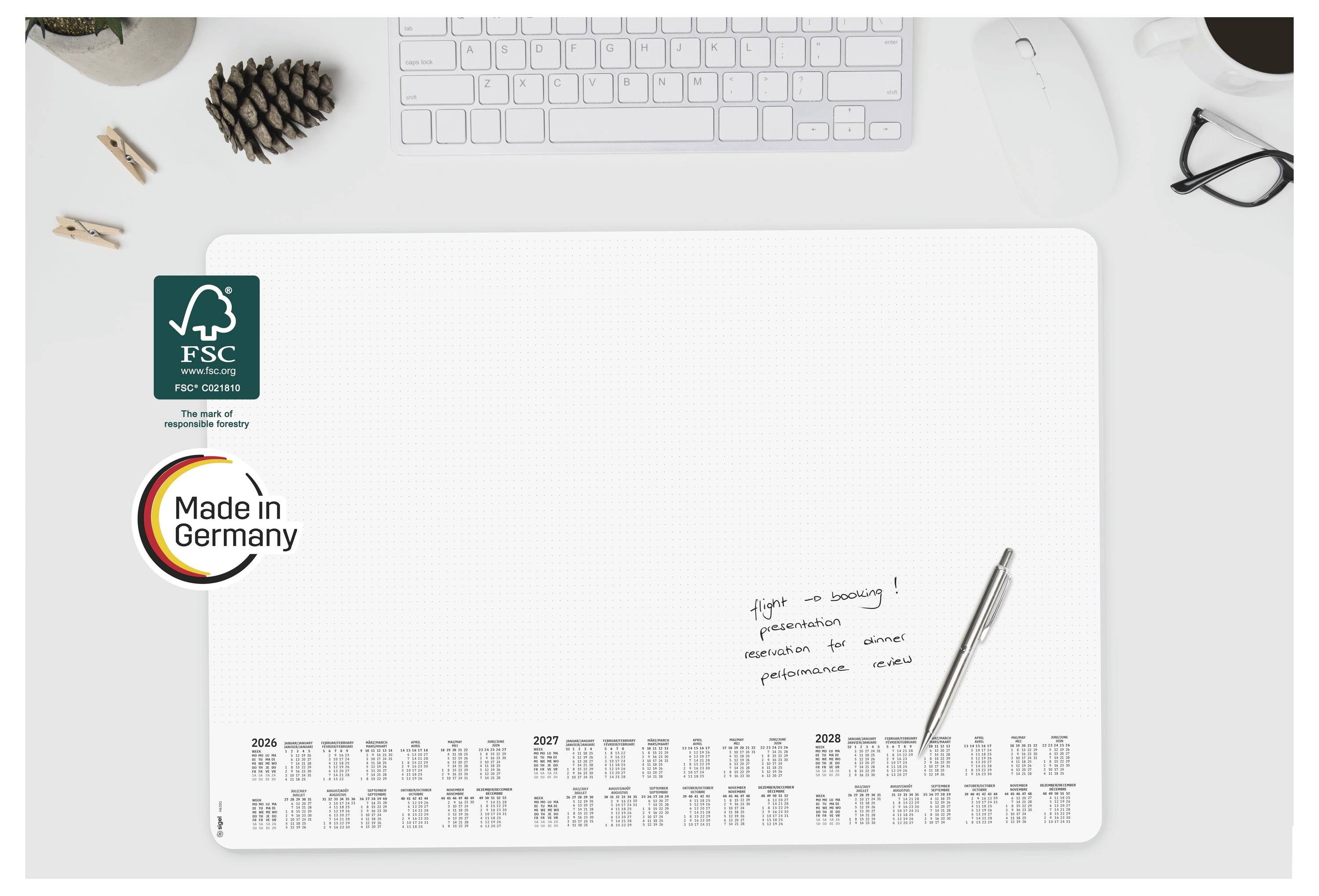 Minimalistischer Schreibtischkalender oder Planer auf weißem Schreibtisch, mit FSC- und „Made in Germany"-Logos gekennzeichnet, mit Tastatur und Stift in der Nähe.
