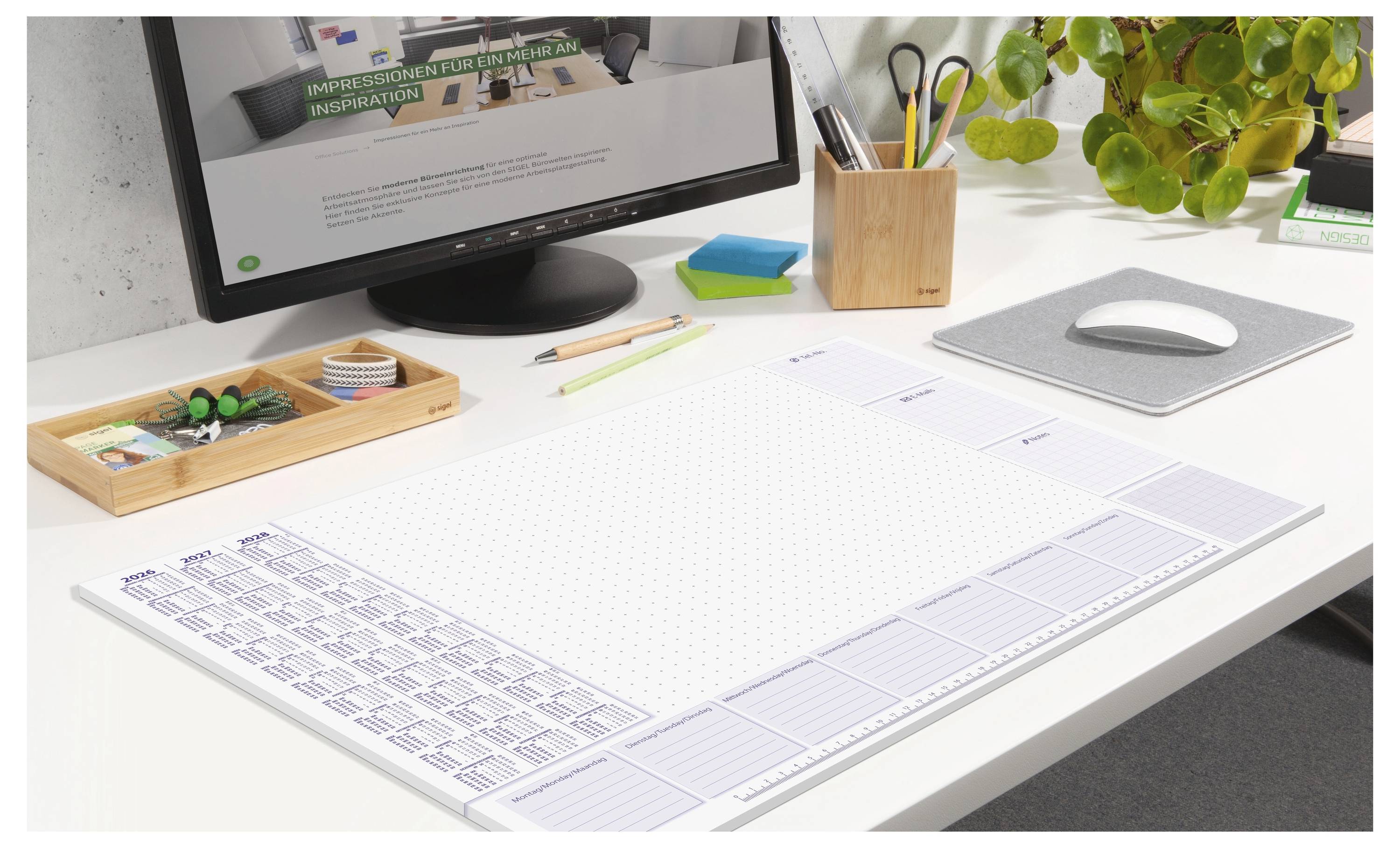 Ein aufgeräumter Schreibtisch mit einem Desktopmonitor, einem Schreibtischkalender, einer Maus auf einer Matte, Stiften in einem Halter und einer kleinen Pflanze; ein Arbeitsplatz-Setup.