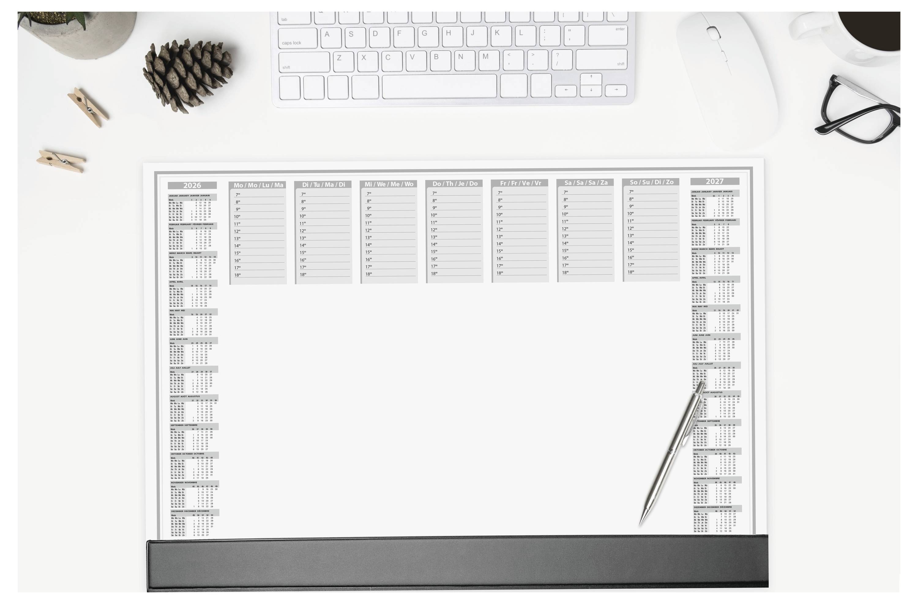 Ein großer Schreibtischkalender liegt auf einer weißen Oberfläche, mit einem Stift darauf. Eine Tastatur, Brille, Tannenzapfen und eine Tasse sind in der Nähe platziert.