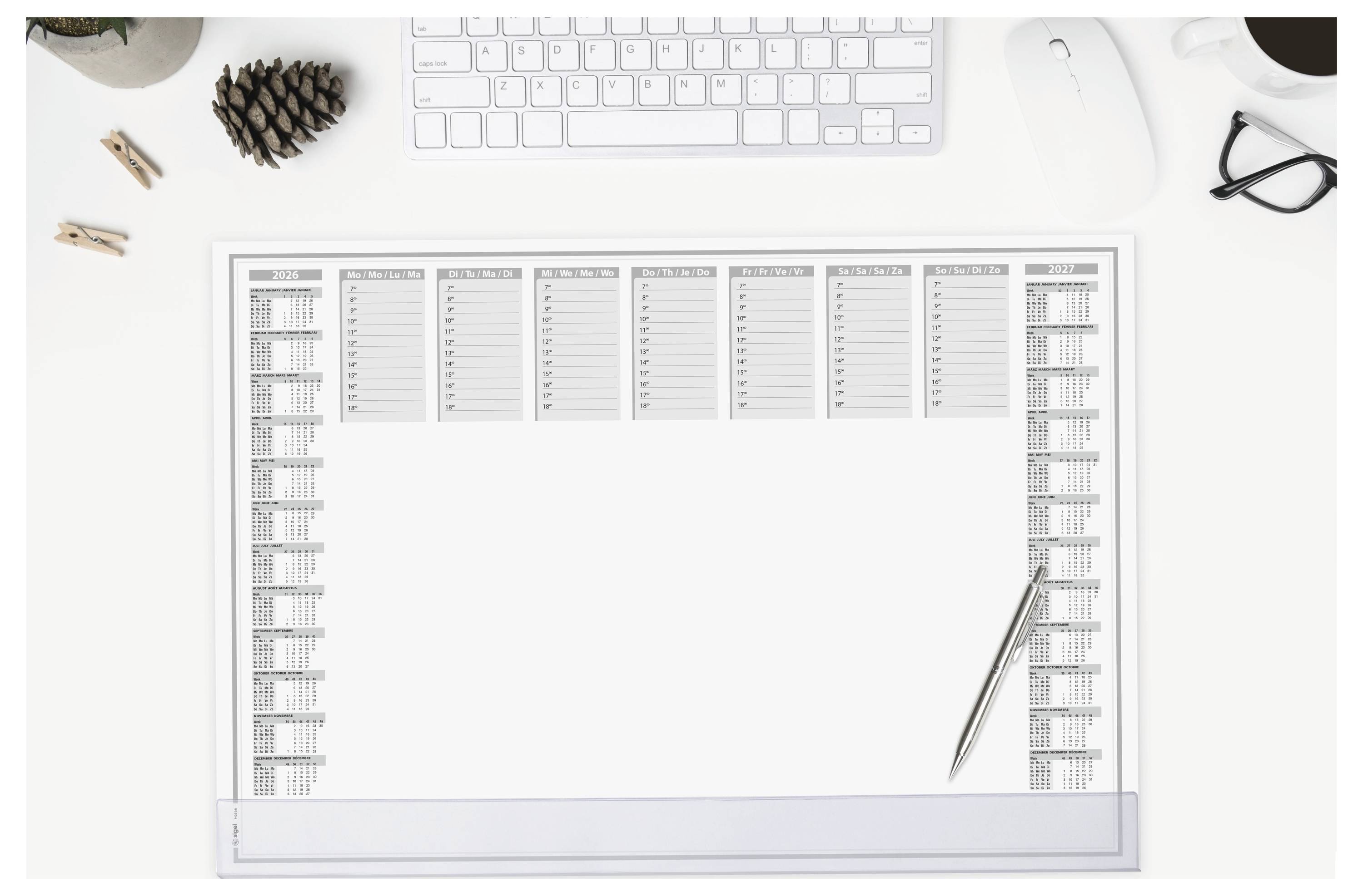 Eine Schreibtischeinrichtung mit einer Tastatur, Maus, Brille, Tannenzapfen und einem Kalenderblock mit einem darauf liegenden Stift.
