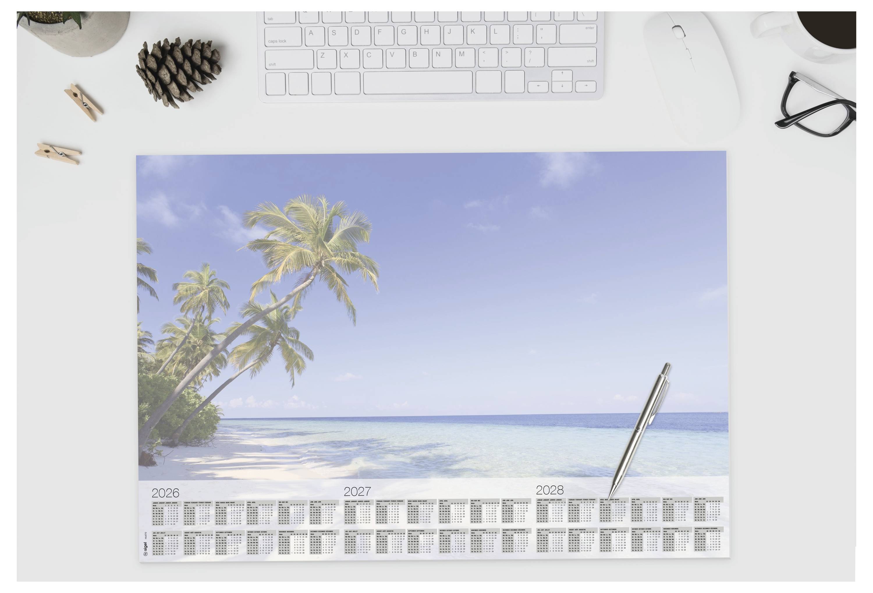 Strandthemen-Schreibtischkalender auf einem weißen Schreibtisch, zeigt eine tropische Küstenlinie mit Palmen, blauem Meer und einem Stift, der auf der Seite ruht.