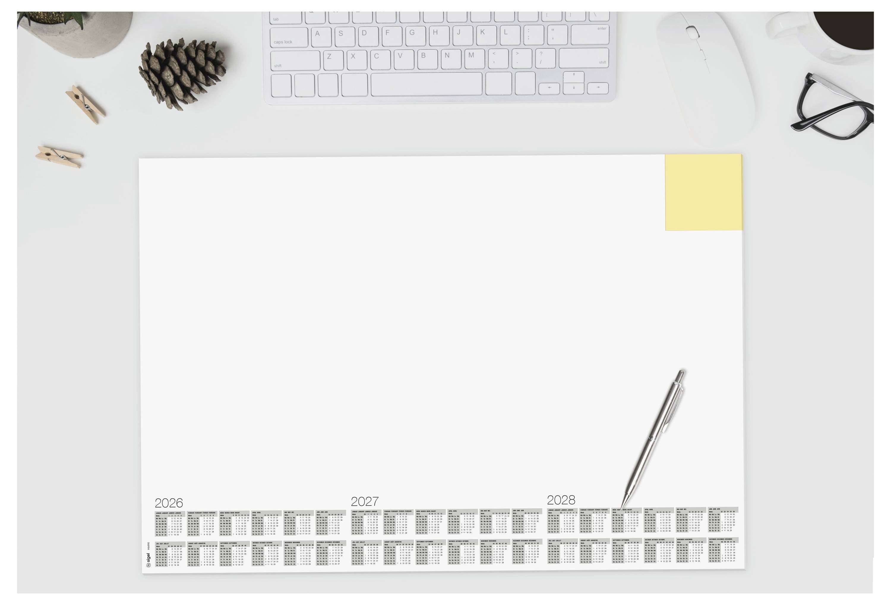 Ein minimalistischer Schreibtisch mit einer Tastatur, einer Tasse, einer Maus und einem Planer mit Jahreskalendern für 2026-2028 und einem Stift obenauf.