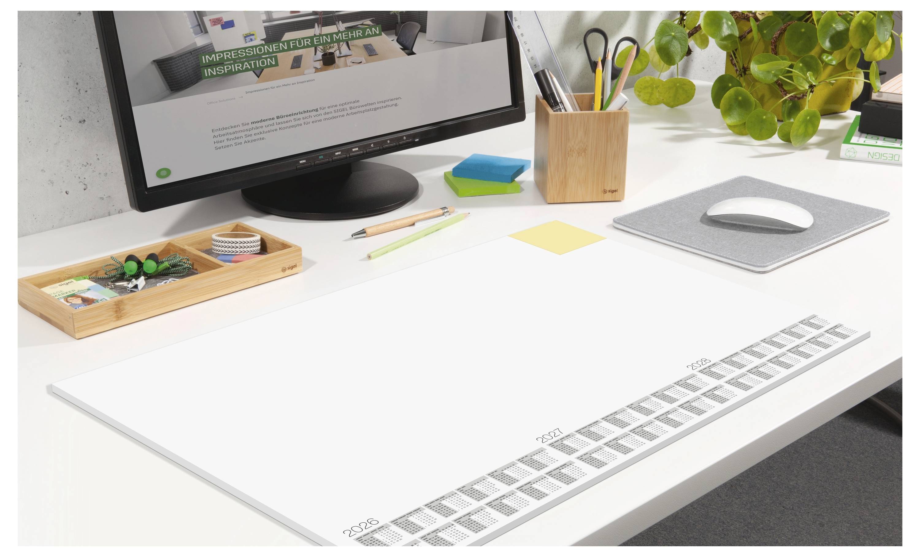 Ein aufgeräumter Schreibtisch mit einem Computermonitor, einer Computermaus, einer Pflanze, Stiften und einem großen Kalenderblock, der Termine am unteren Rand anzeigt.